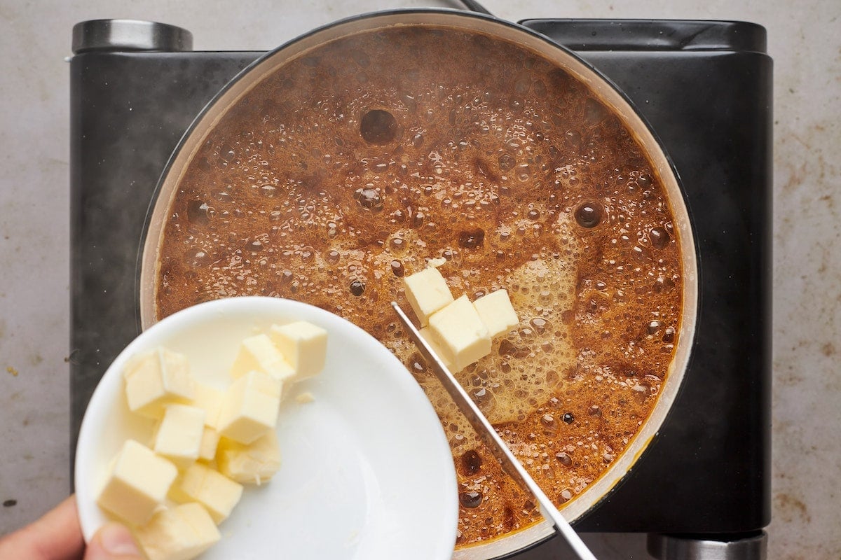 adding butter pieces to boiling salted caramel sauce in pan. 