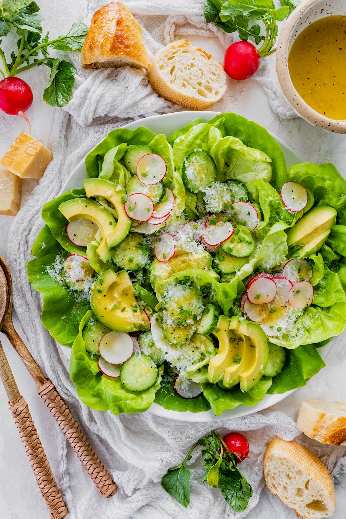 butter lettuce salad with avocado, cucumber, radish slices, chives, Parmesan cheese, and lemon dressing In large white bowl with baguette on the side. 