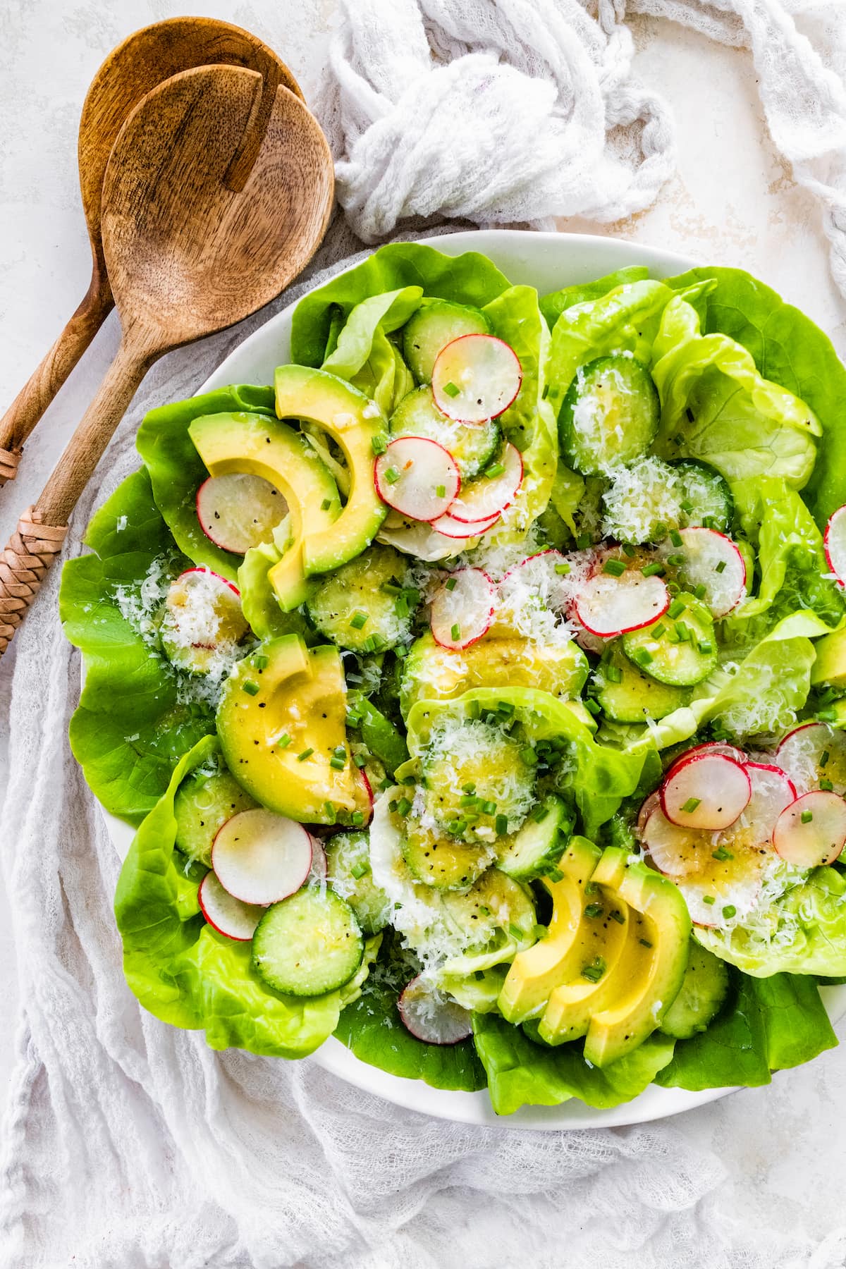 butter lettuce salad with avocado, cucumber, radish slices, chives, Parmesan cheese, and lemon dressing In large white bowl with wood salad servers. 
