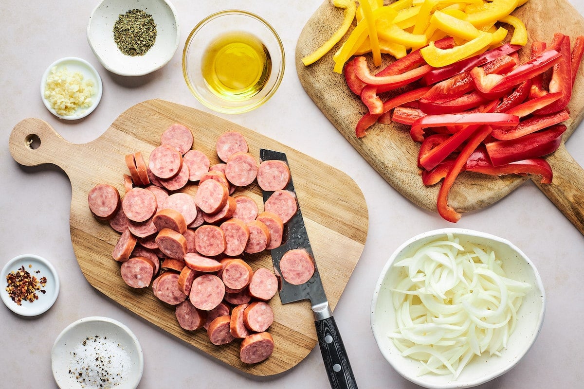 sliced sausage on wood cutting board with knife, bowl of onions, bell pepper strips on wood cutting board, and small bowls with olive oil, salt, pepper, garlic, and crushed red pepper flakes. 