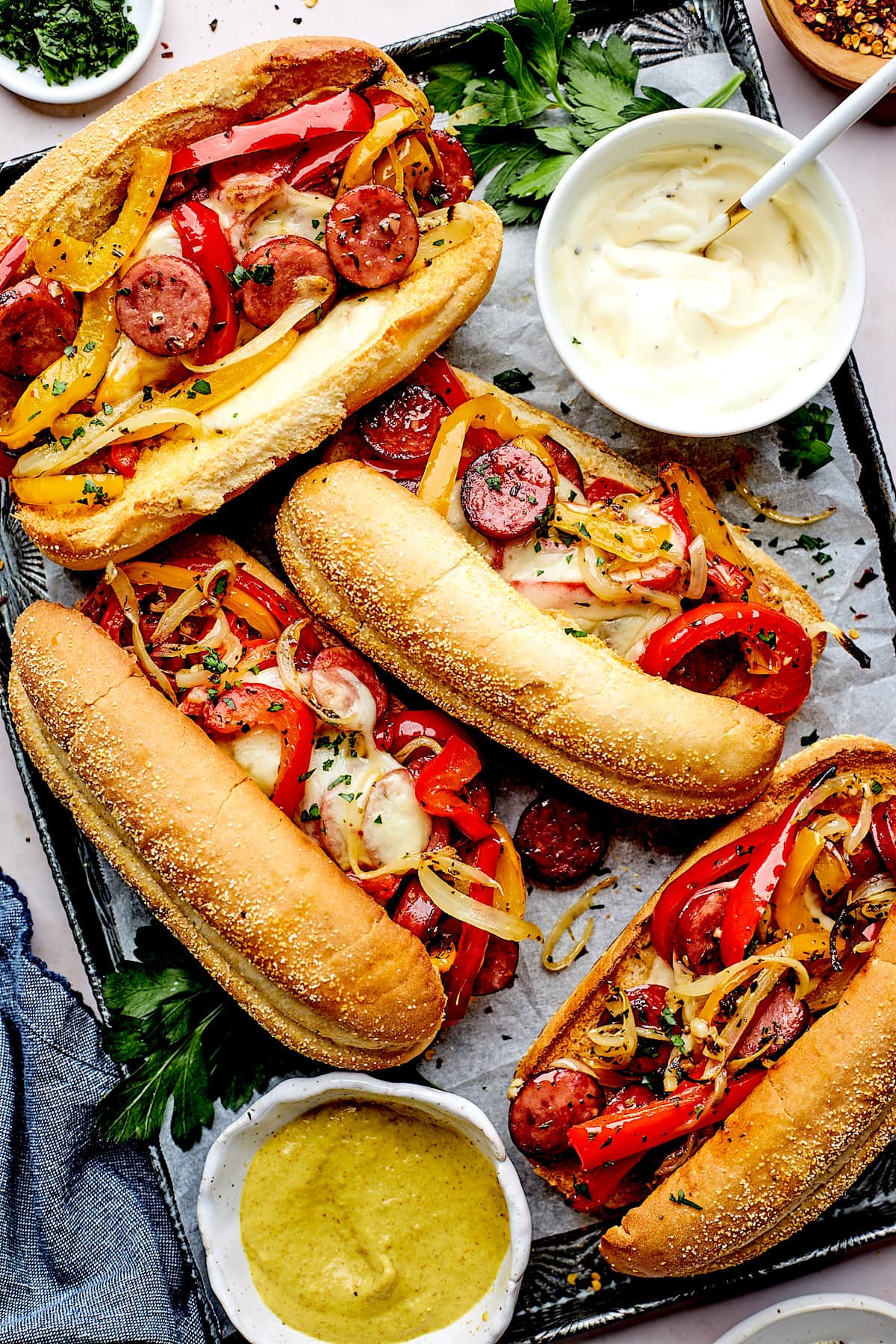 sausage and peppers hoagie sandwiches with melted provolone cheese on sheet pan with mustard and mayonnaise in bowls. 