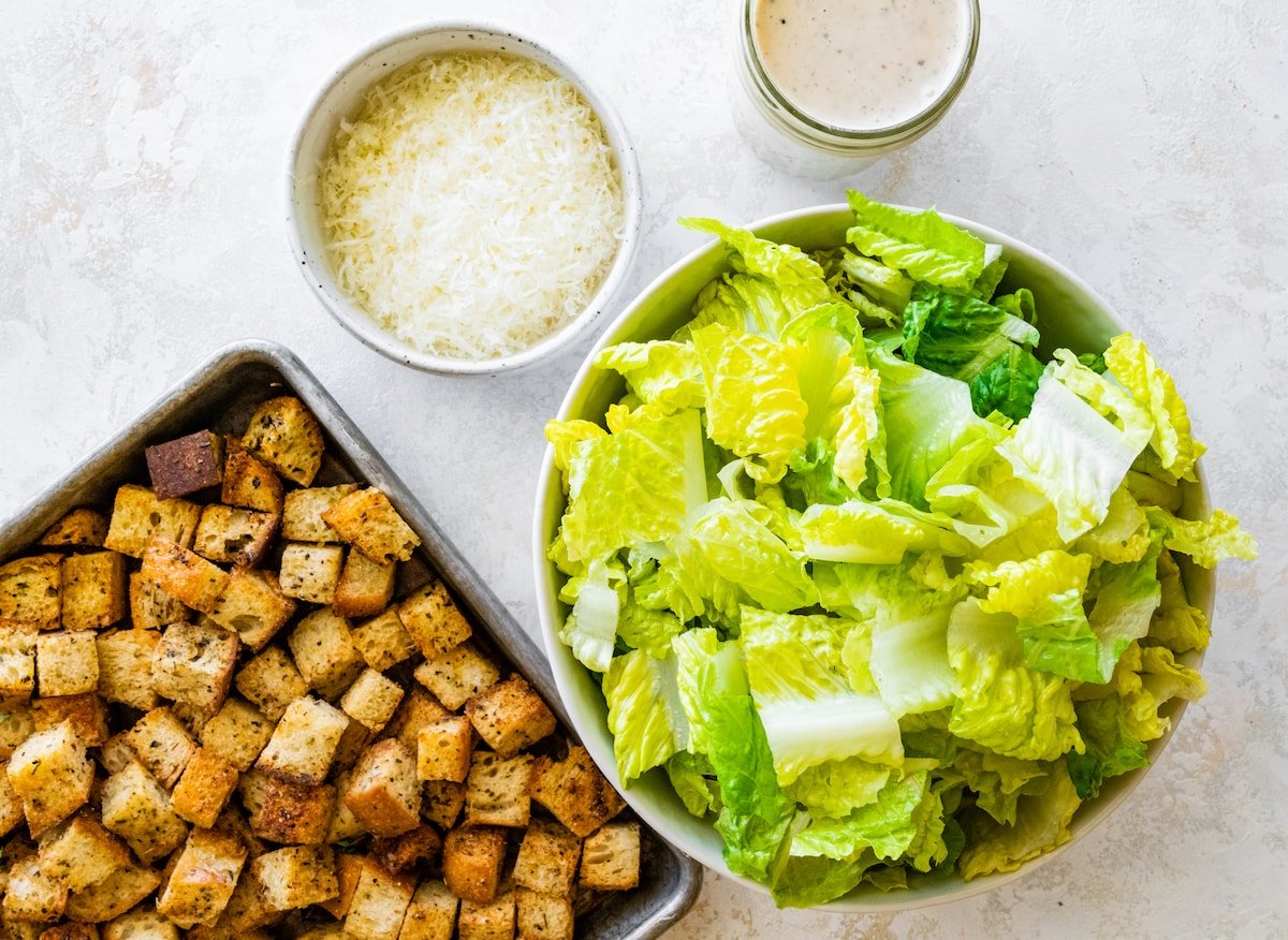 homemade croutons on baking sheet, romaine lettuce in bowl, Parmesan cheese in bowl, and Caesar dressing. 