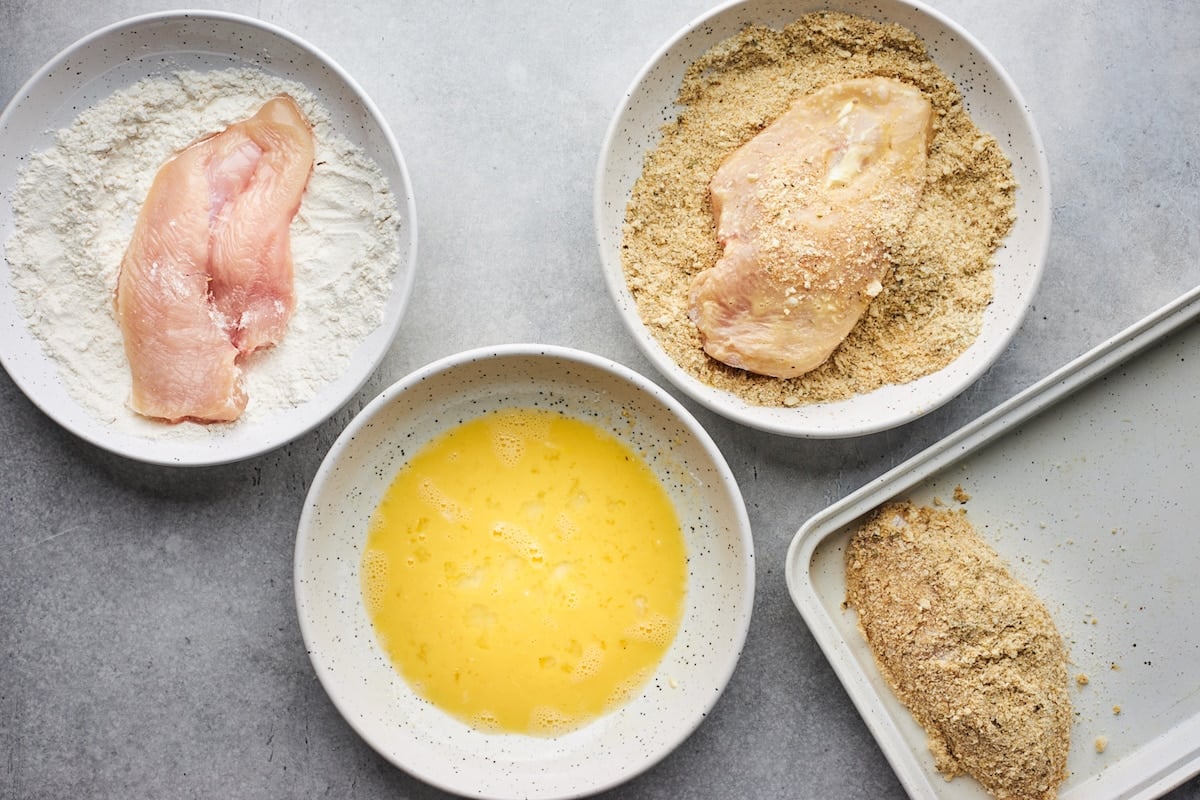 chicken breast in bowl of flour with a bowl of beaten eggs and chicken breast in bowl of breadcrumbs to make chicken parmesan.