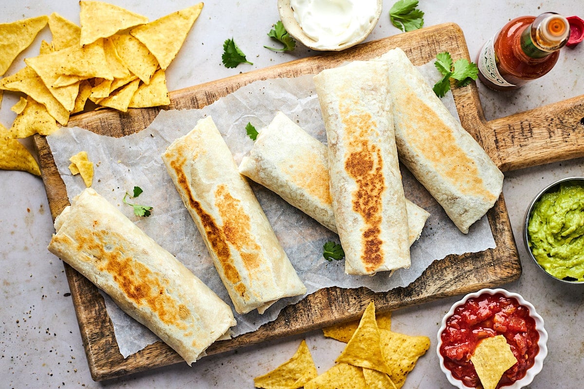 bean and cheese burritos on wood board with chips, salsa, and guacamole. 