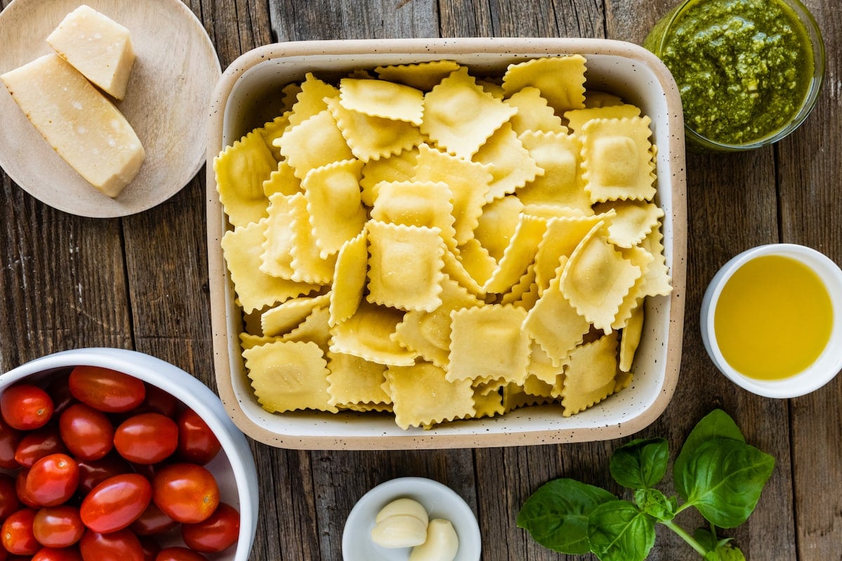 cheese ravioli in dish, tomatoes in bowl, Parmesan cheese on plate, pesto in bowl, olive oil in bowl, basil, and garlic. 