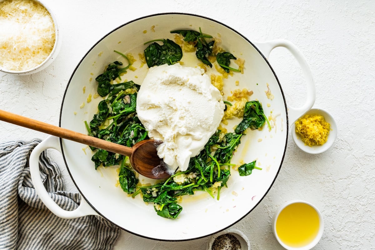 wilted spinach, spinach, garlic, and ricotta cheese in white pot with wooden spoon. 