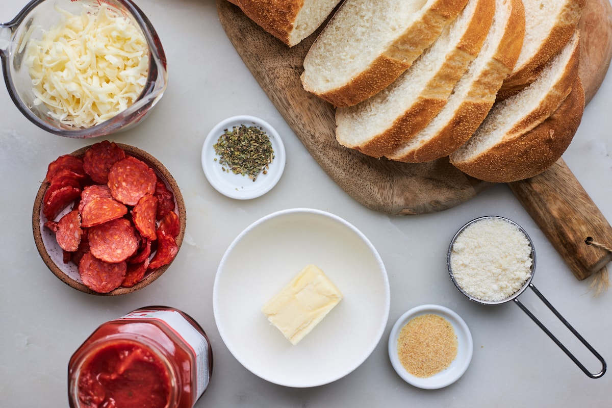 sliced bread on a wood board, shredded mozzarella cheese, pepperoni, pizza sauce, butter, garlic, Italian seasoning, and Parmesan cheese to make pizza grilled cheese.