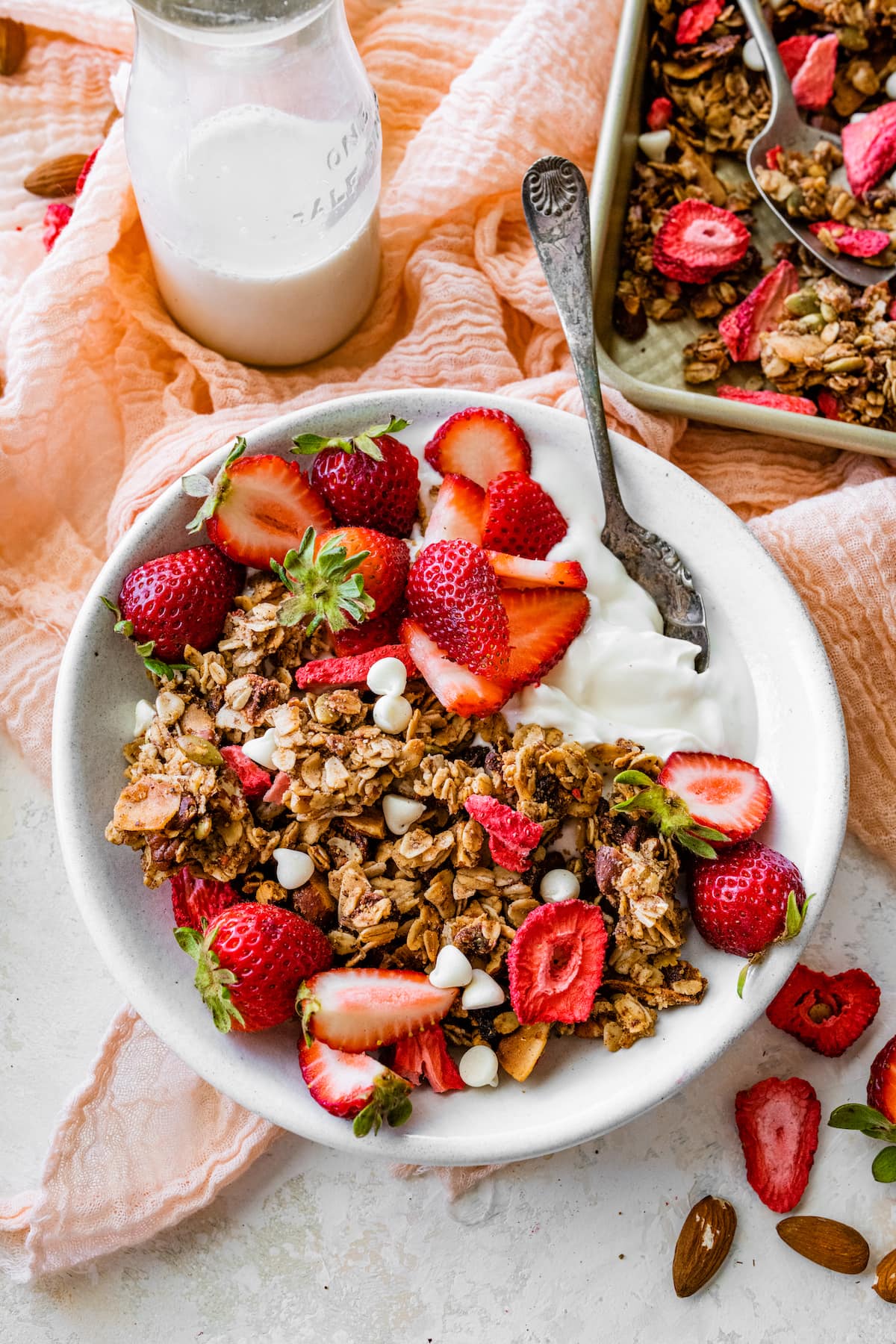 strawberry granola in bowl with yogurt, fresh strawberries, white chocolate chips, and spoon. 