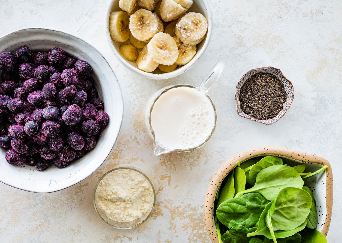 frozen blueberries, frozen banana, milk, protein powder, chia seeds, and spinach in bowls to make a blueberry smoothie. 