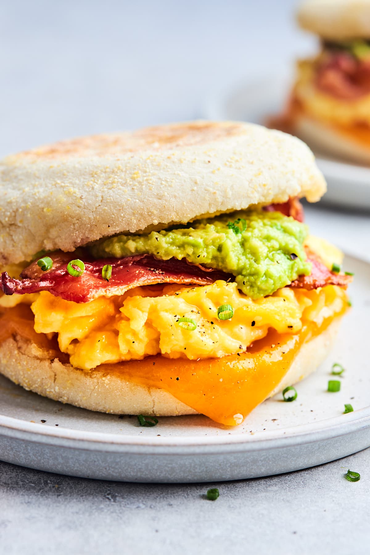 breakfast sandwich with scrambled eggs, cheese, bacon, avocado, and chives on plate. 