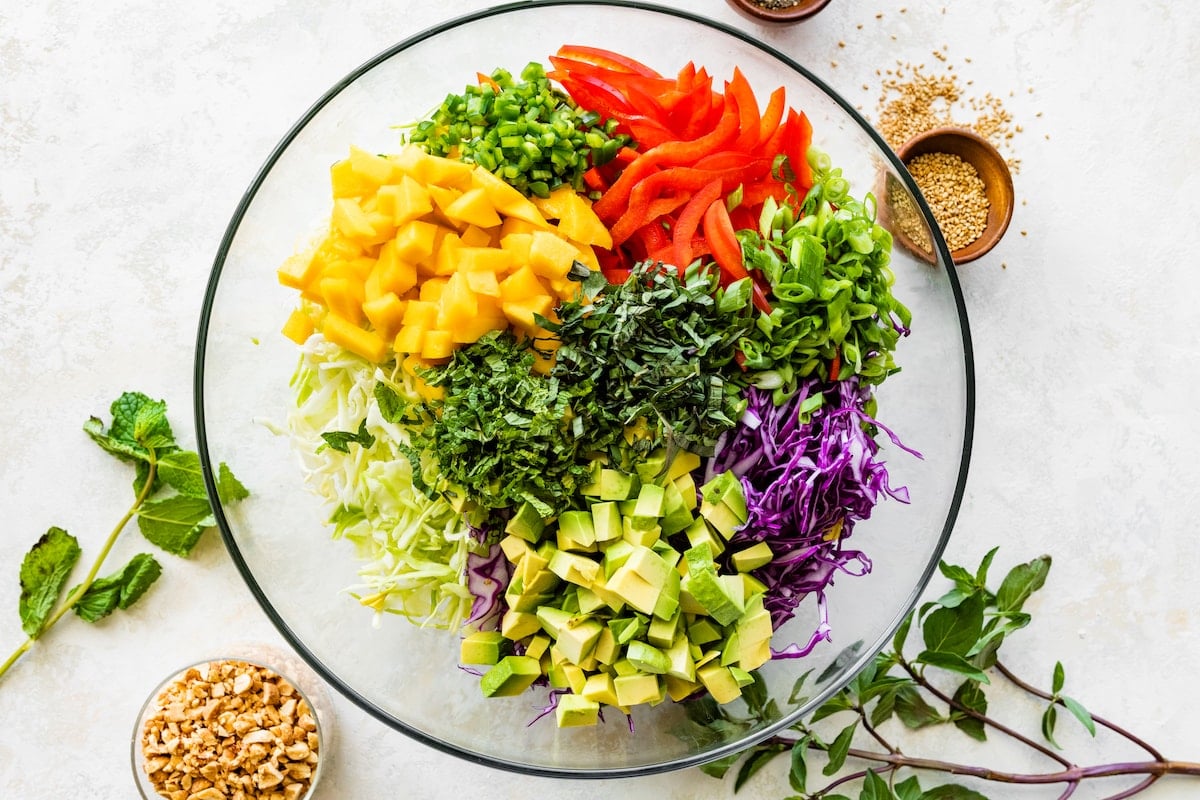 shredded cabbage, mango, red bell pepper, onion, herbs, and avocado in bowl to make cabbage salad. 