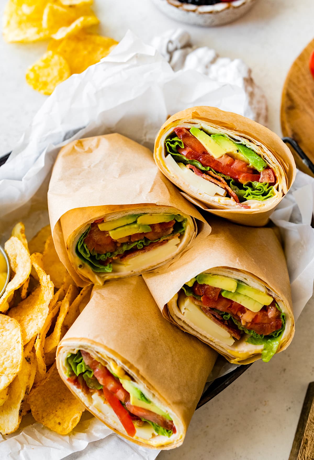 turkey wraps with cheese, bacon, tomato, avocado, and lettuce wrapped in parchment paper in basket with chips. 