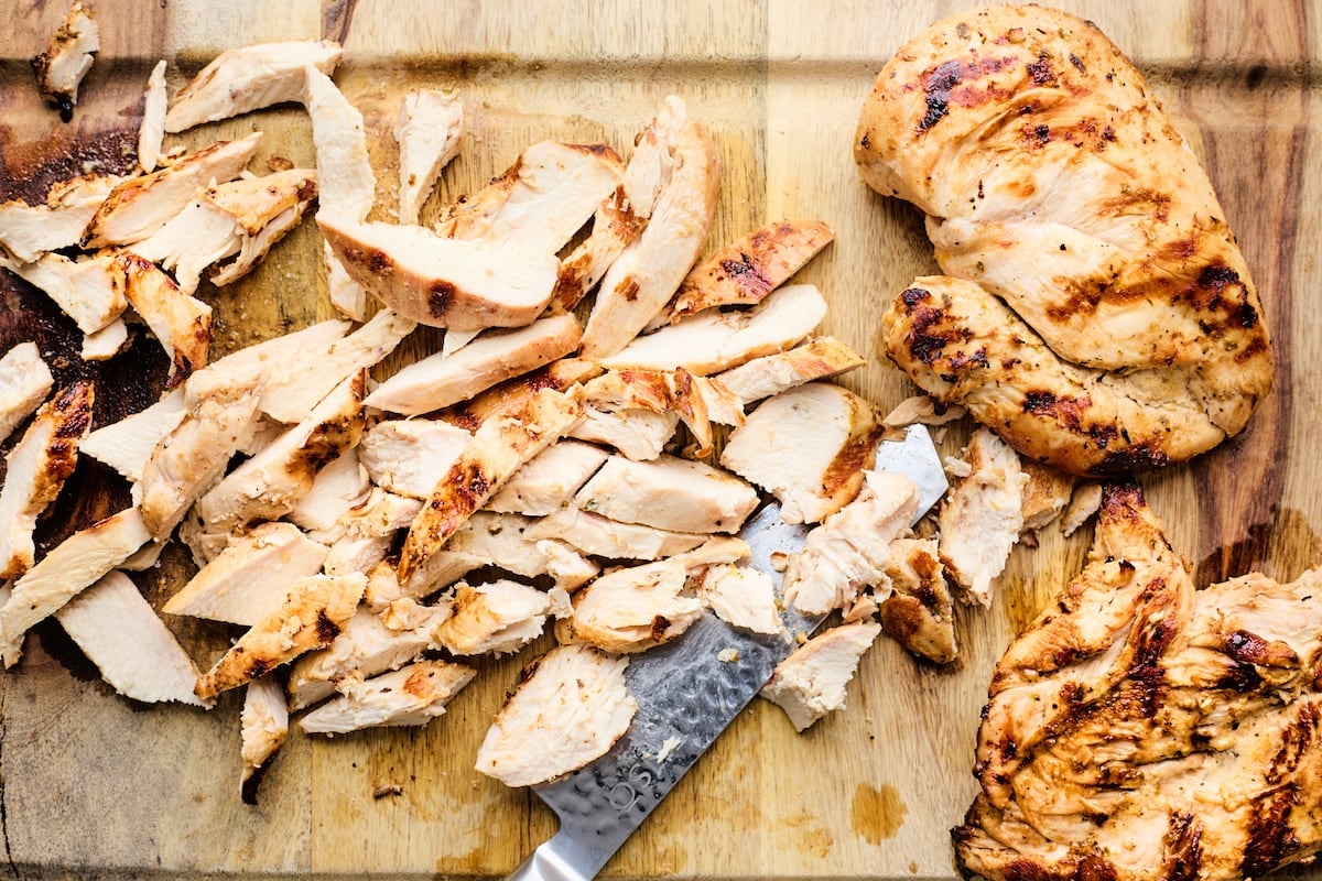 grilled chicken being chopped with sharp knife on cutting board. 