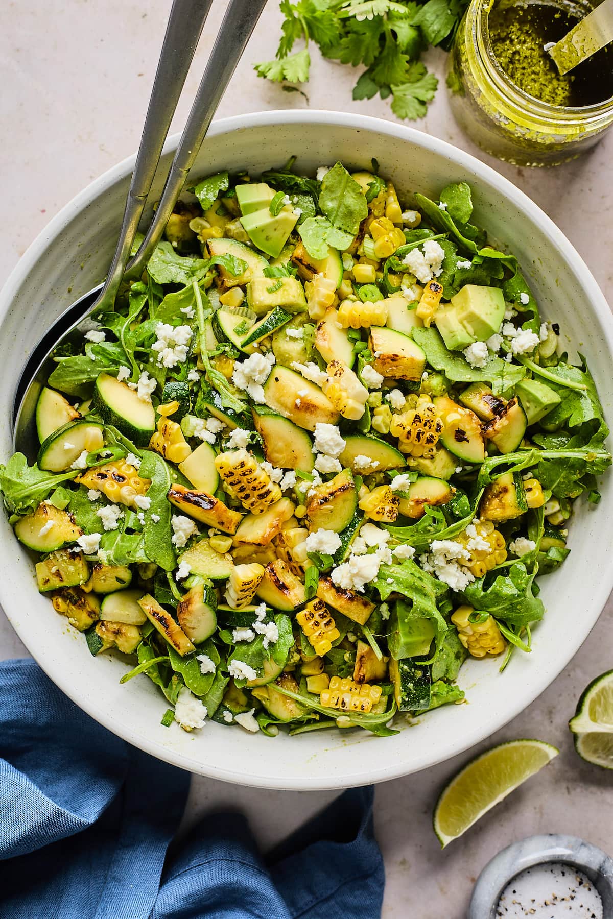 grilled corn zucchini salad with arugula, avocado, queso fresco and cilantro lime vinaigrette in a bowl with serving spoons. 