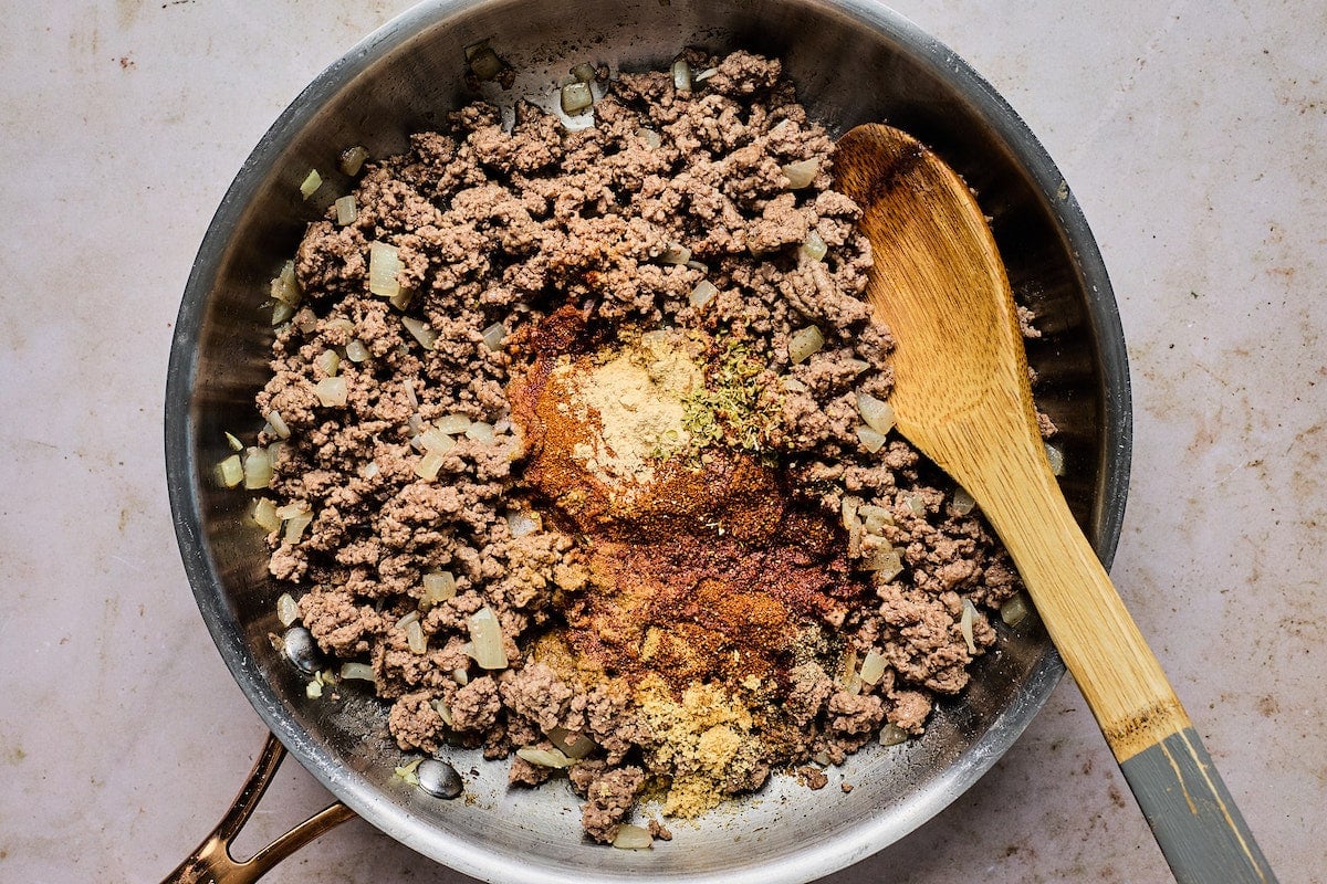 beef, onion, and spices in skillet cooking with wooden spoon. 