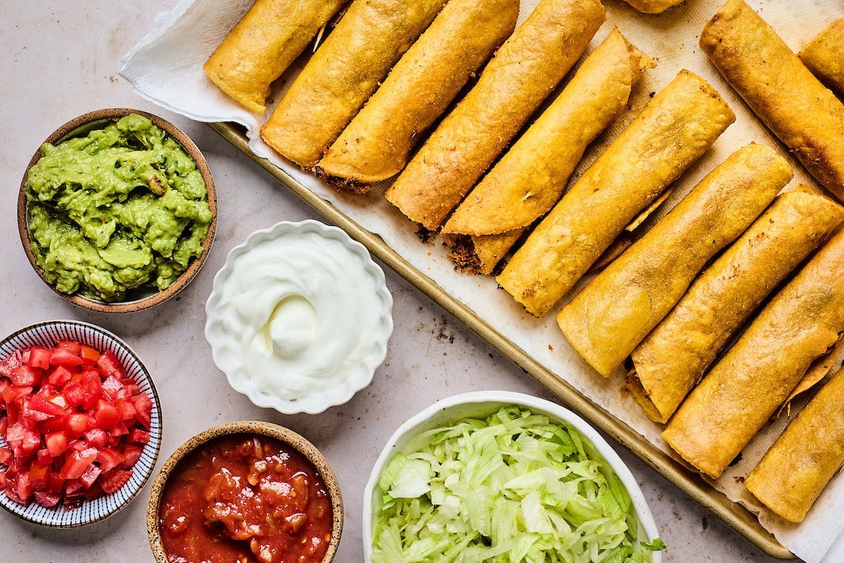 beef taquitos on baking sheet with toppings in bowls. 