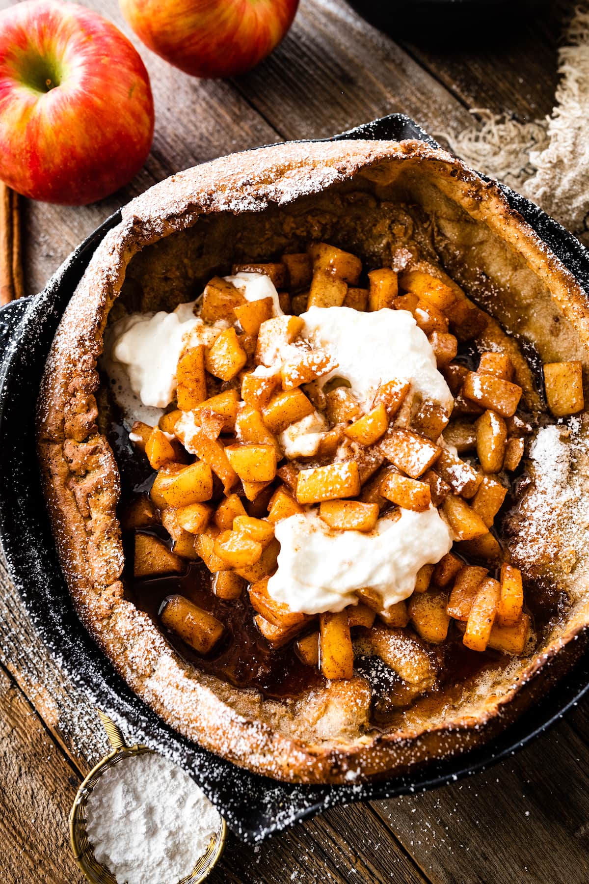 brown butter dutch baby pancake in cast iron skillet with cinnamon apples, whipped cream, and confectioner's sugar. 