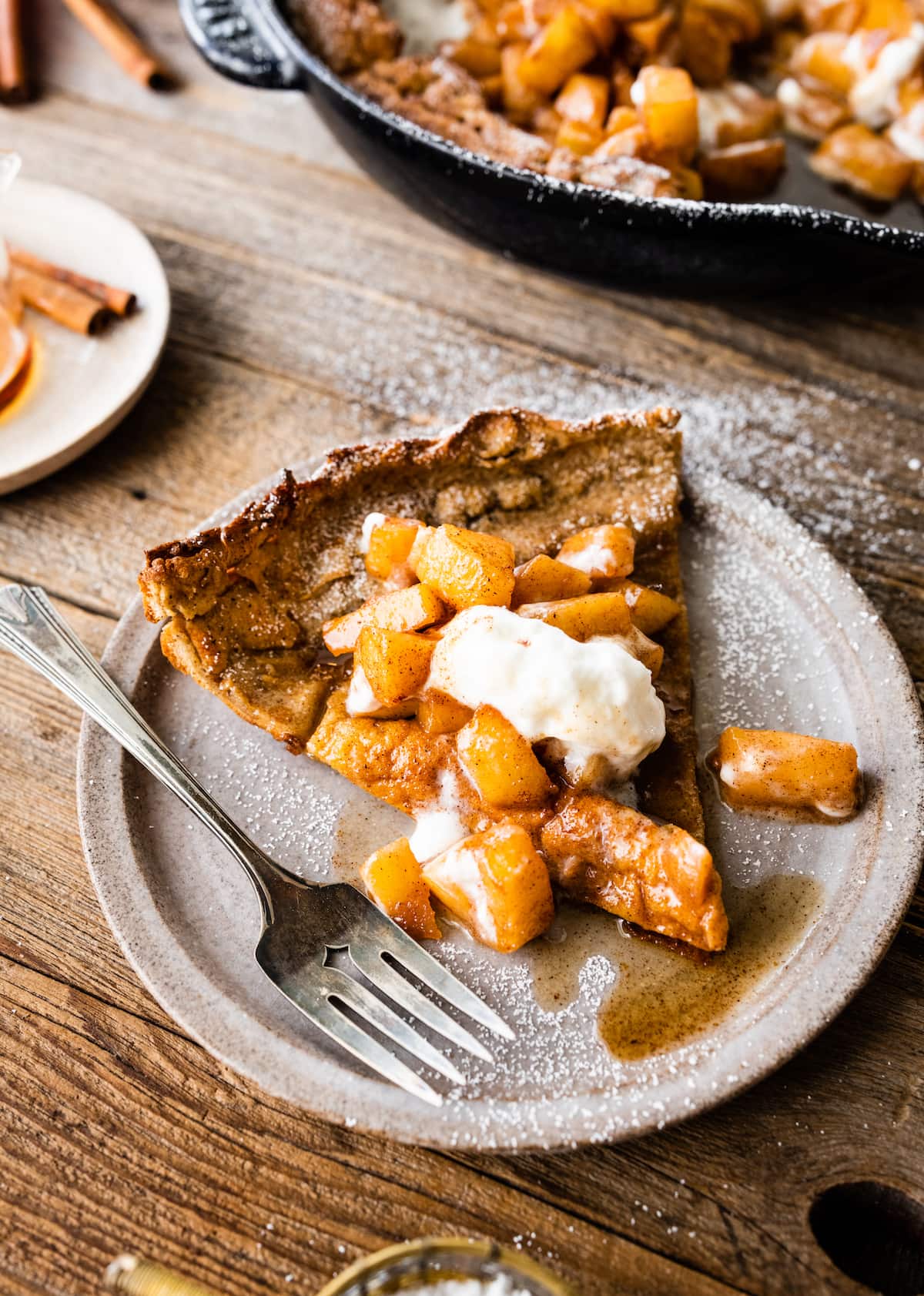 brown butter Dutch baby pancake on plate with cinnamon apples, whipped cream, confectioner's sugar, and maple syrup. 