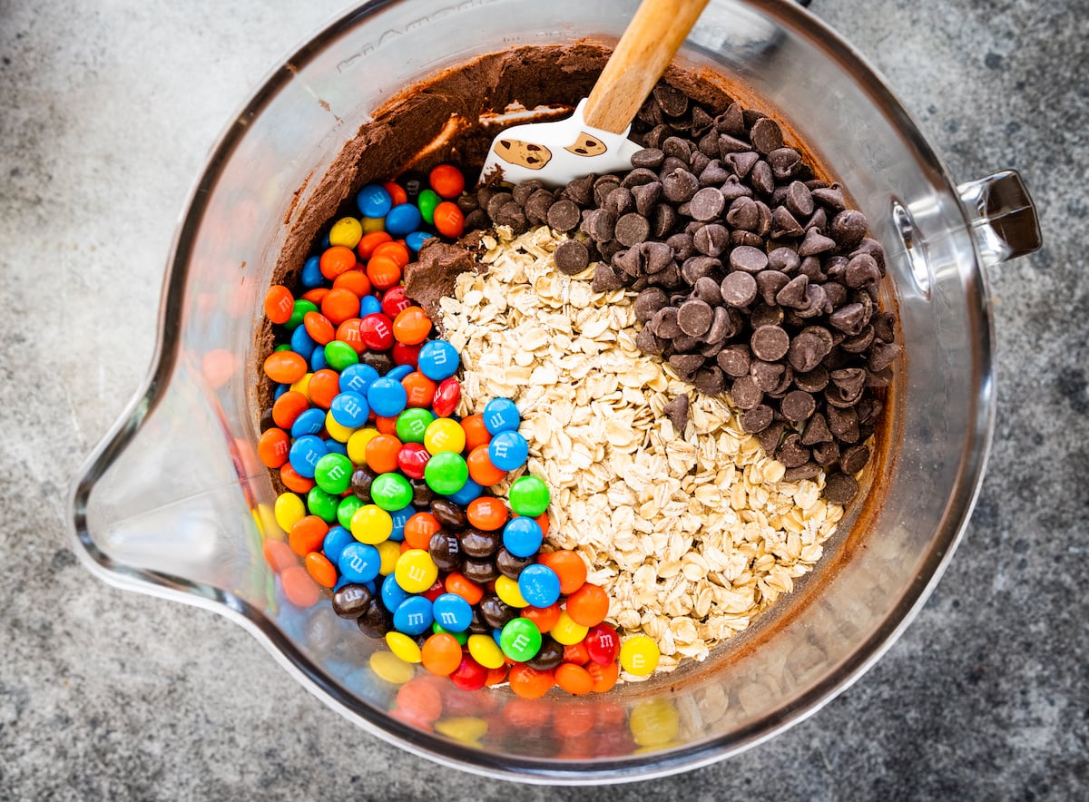 M&M's, oats, and chocolate chips being stirred into monster cookie dough with spatula. 