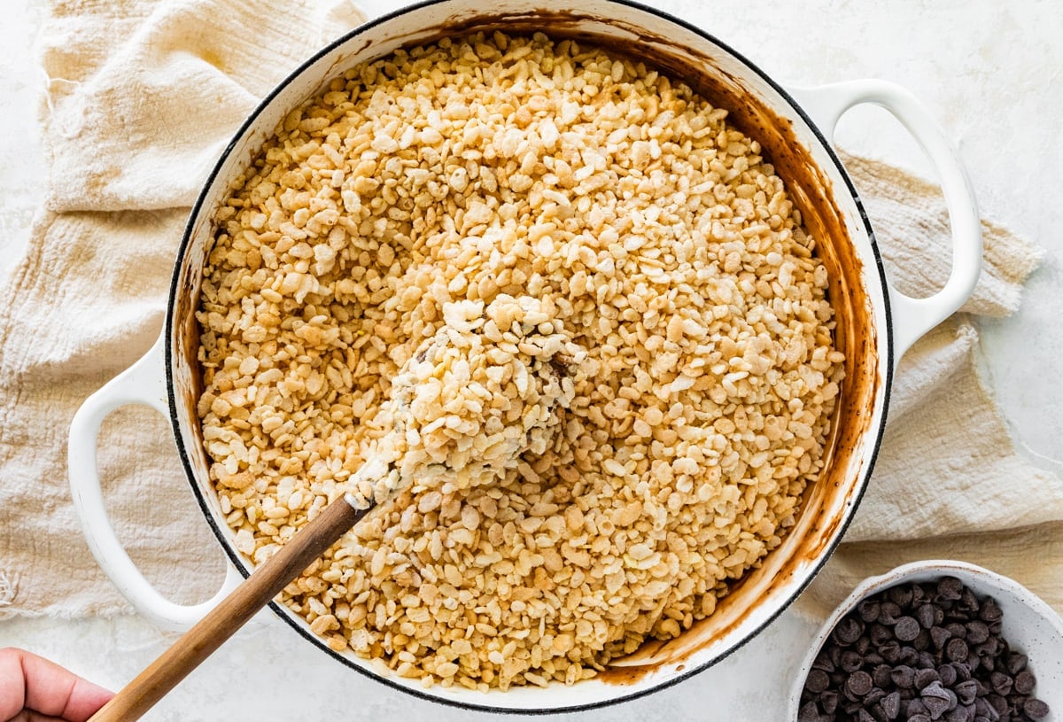 crispy rice cereal being stirred into melted marshmallows in pot with wooden spoon. 