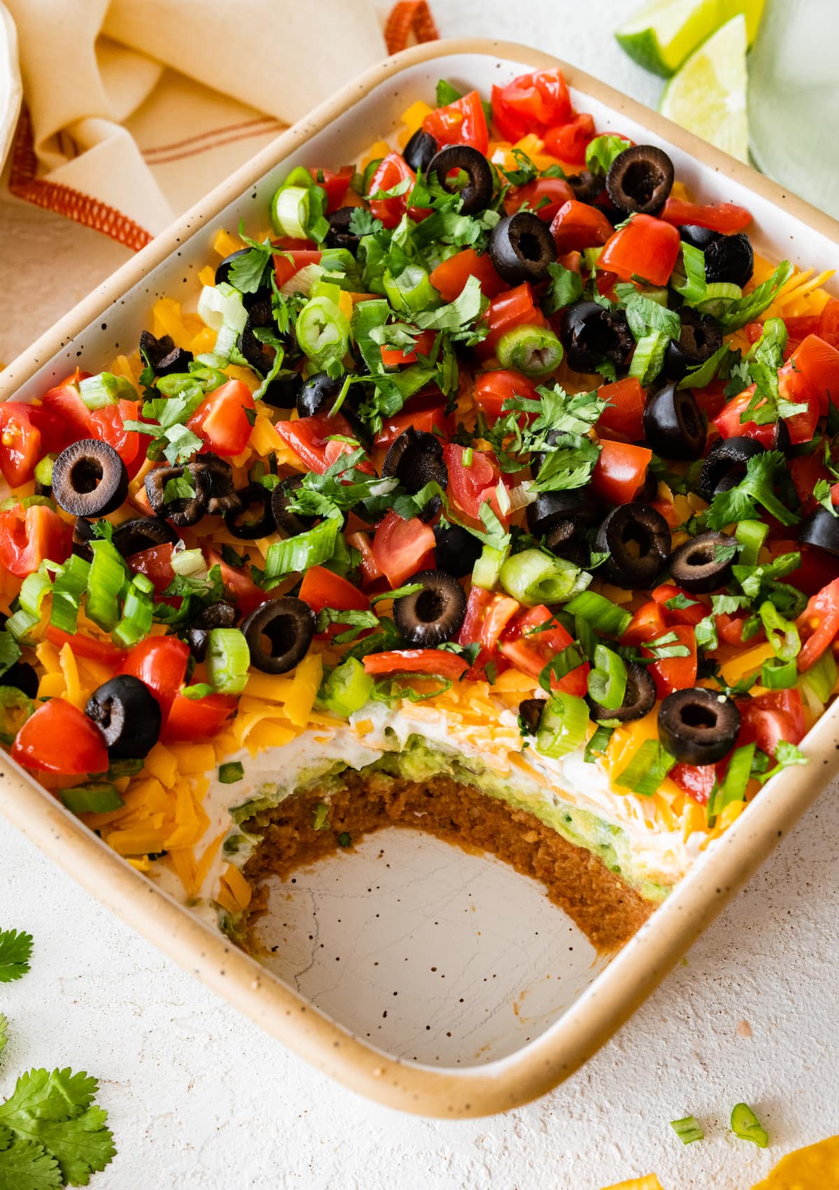 7 layer dip with a layer of refried beans, guacamole, sour cream, cheese, tomatoes, black olives, green onions, and cilantro in baking dish. 