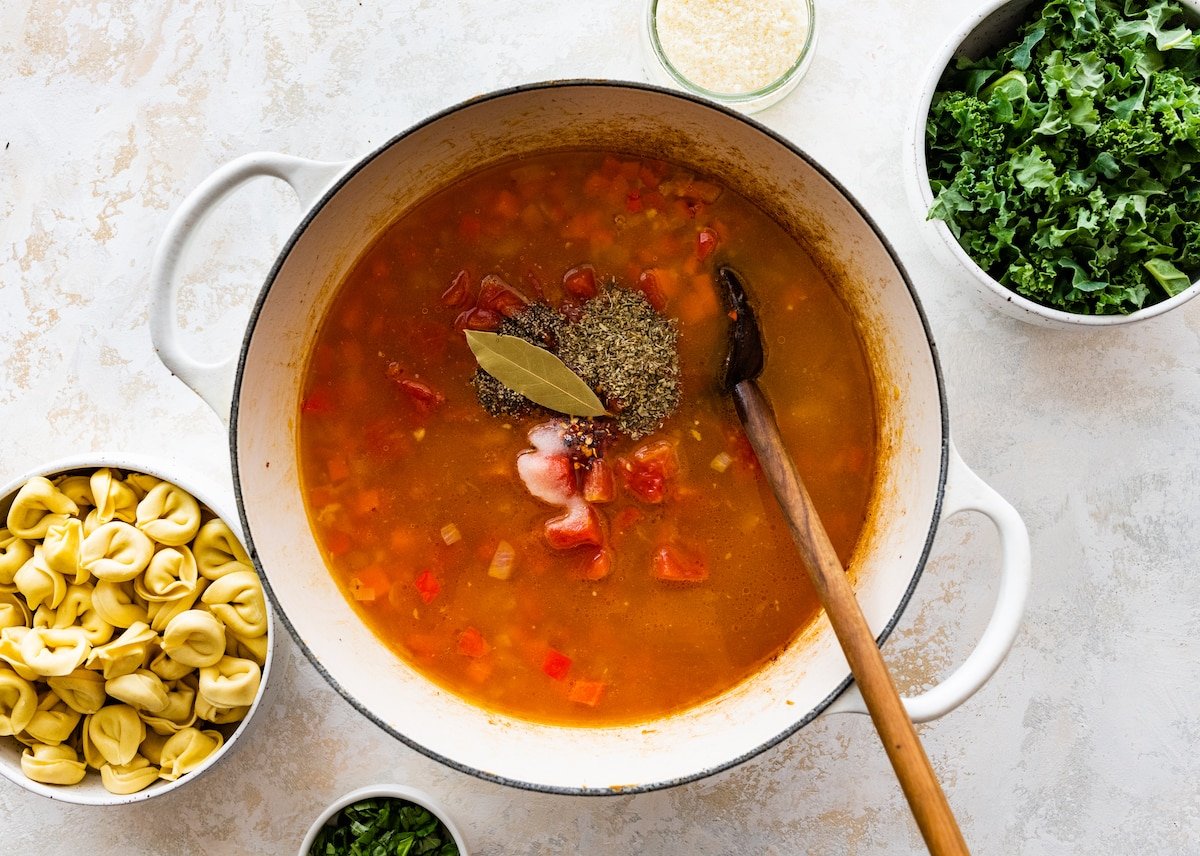 veggies, broth, herbs, and spices in pot with wooden spoon to make tortellini soup. 