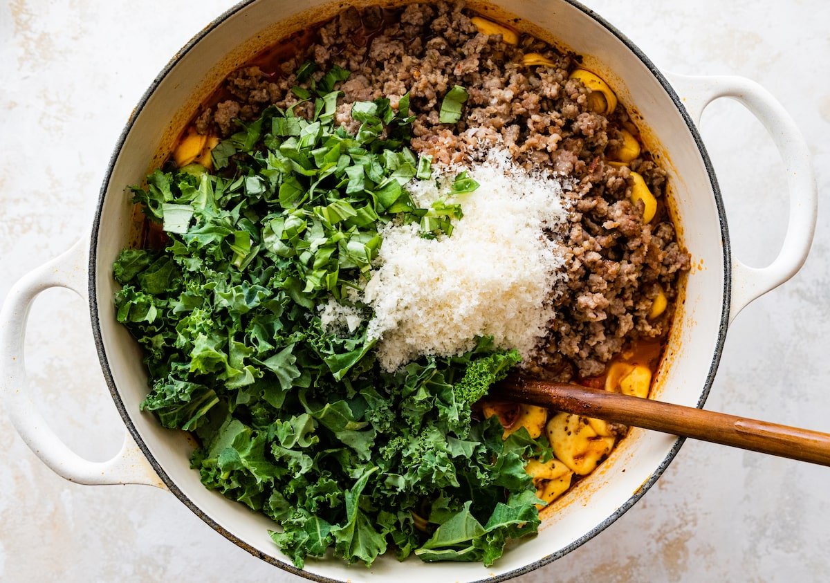 stirring in sausage, kale, and Parmesan cheese to tortellini soup in pot. 