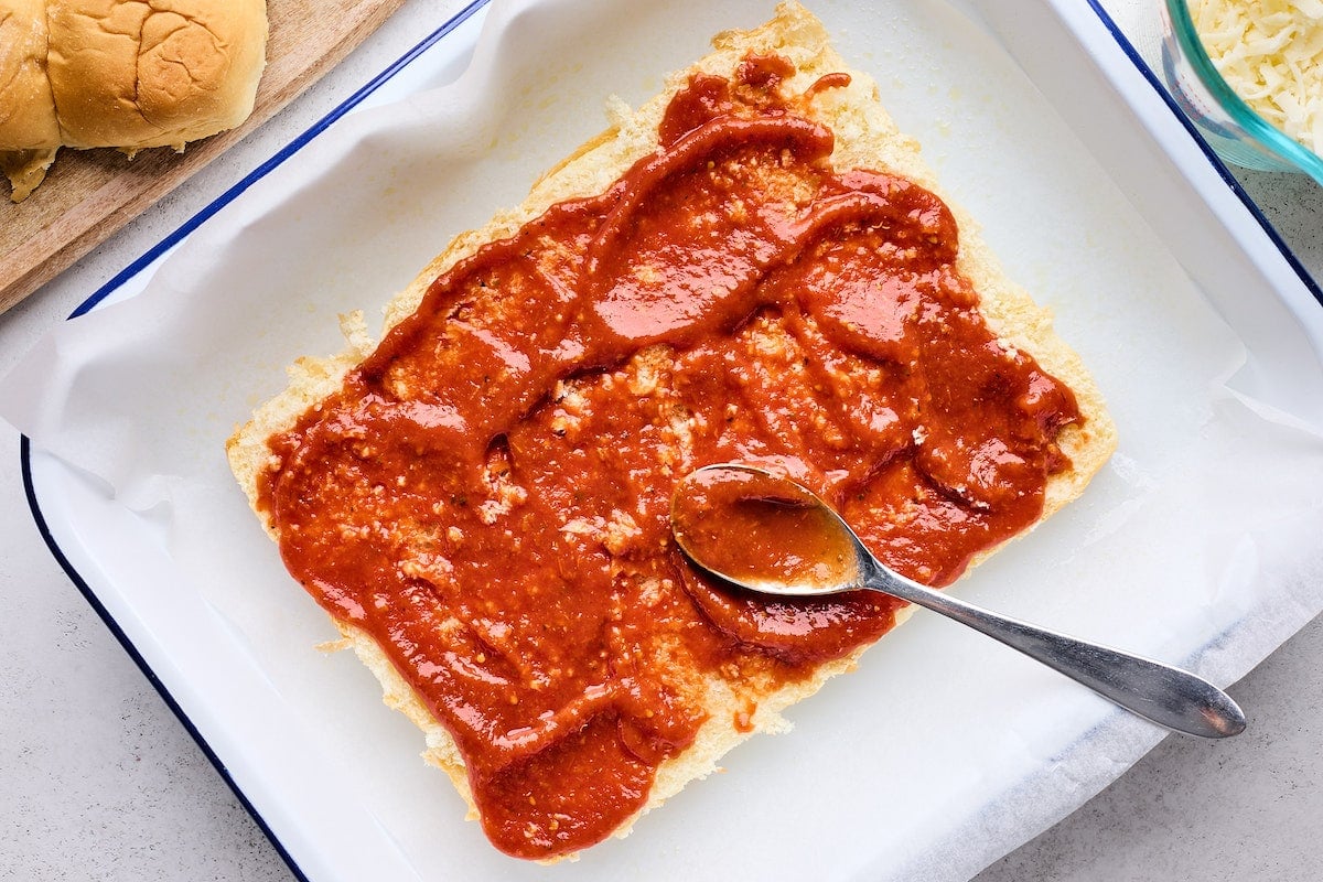 pizza sauce being spread on Hawaiian sweet rolls with spoon. 