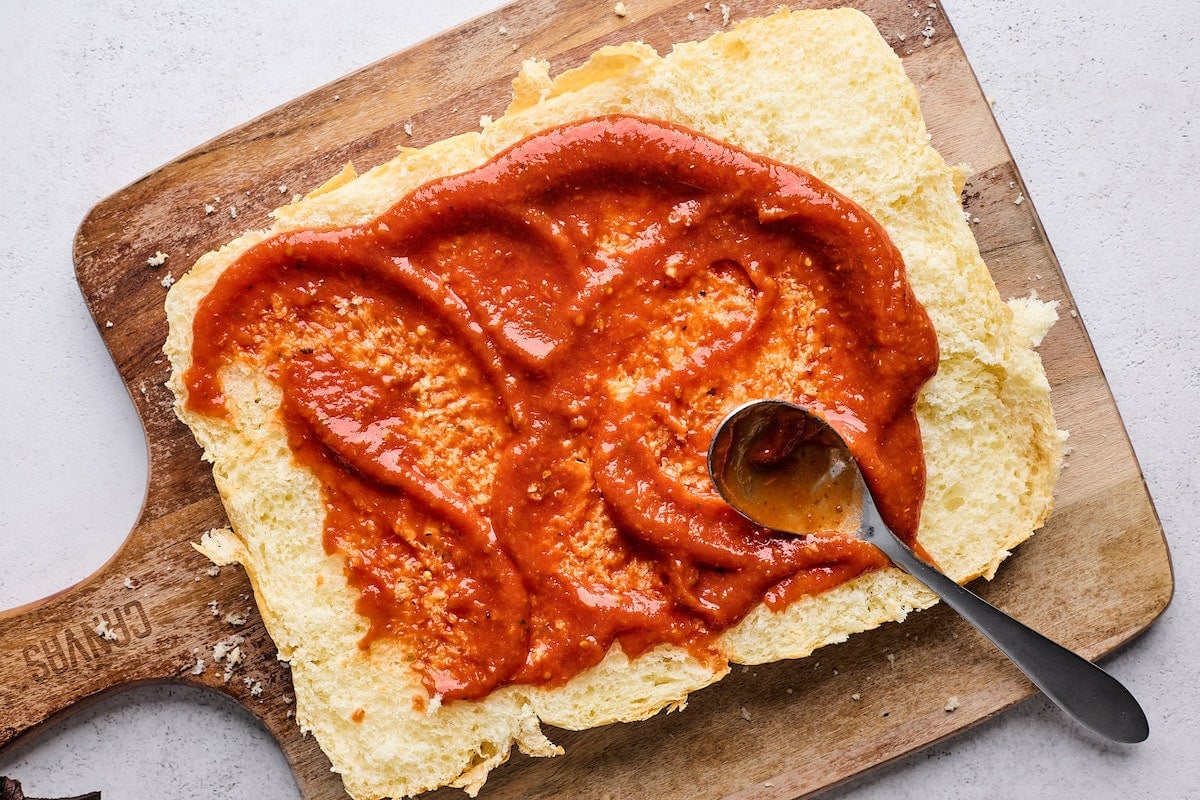 pizza sauce being spread on top layer of Hawaiian sweet rolls with spoon. 