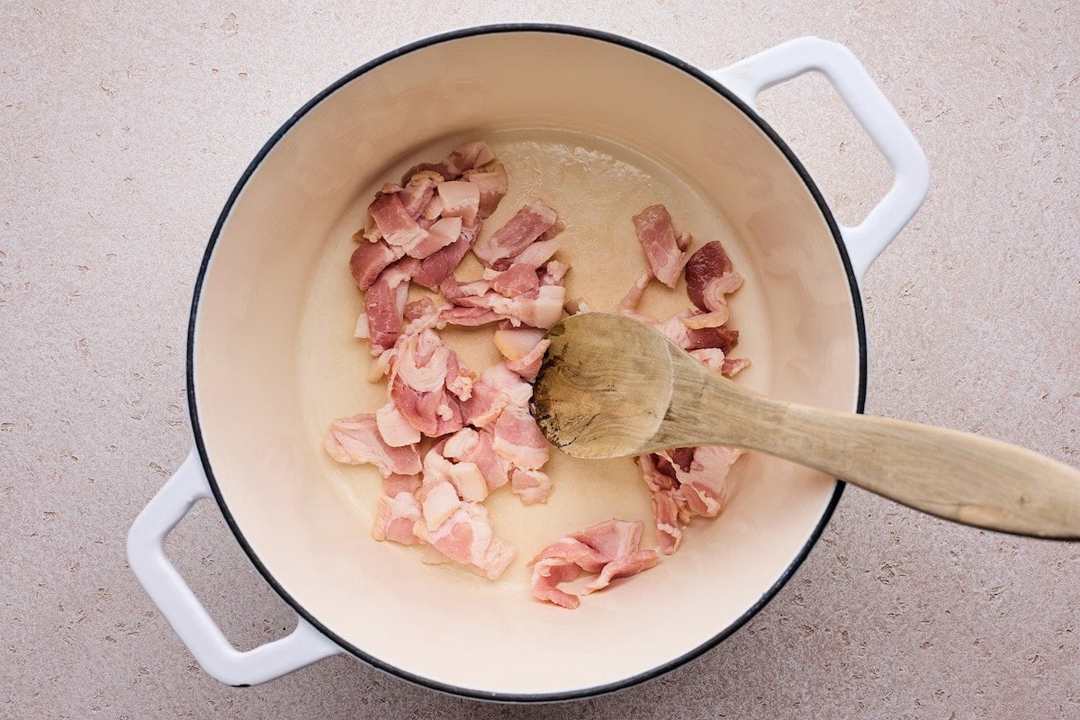 bacon cooking in white pot with wooden spoon to make zuppa toscana. 