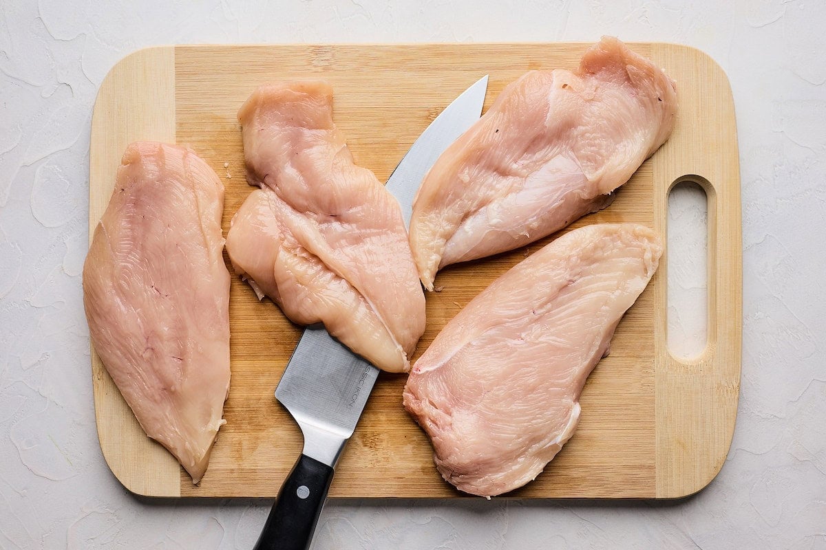 chicken breasts being cut in half with a sharp knife on a cutting board. 