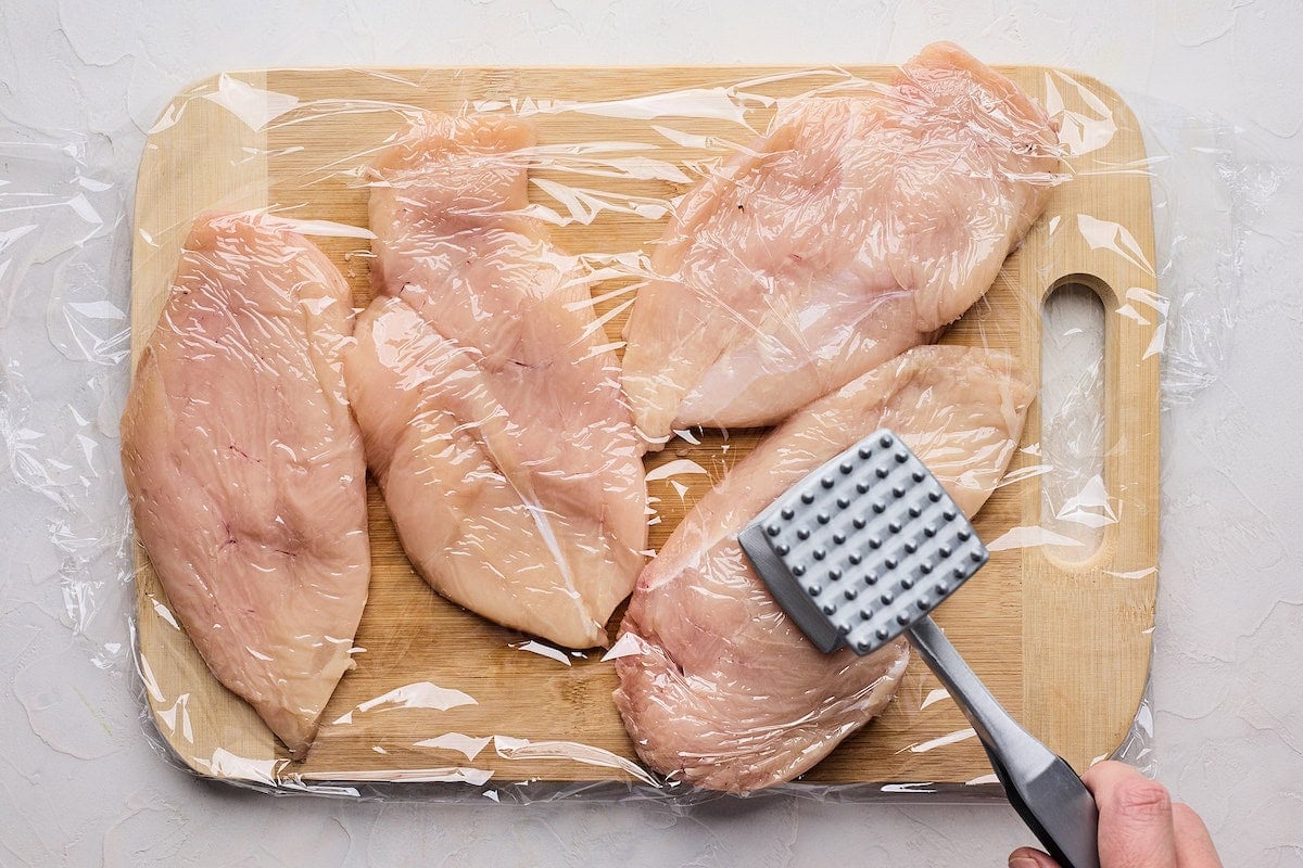 chicken breasts being pounded with meat mallet on cutting board. 