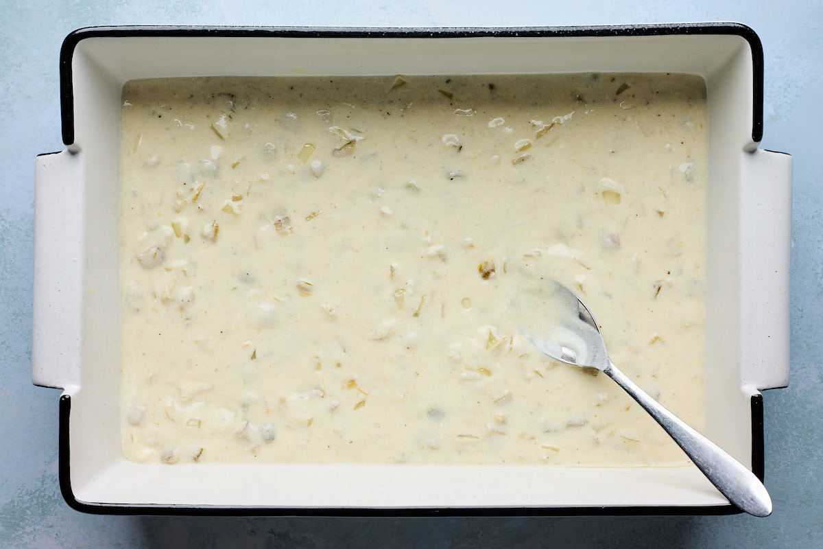 creamy white chicken enchilada sauce being spread in bottom of baking dish with spoon. 