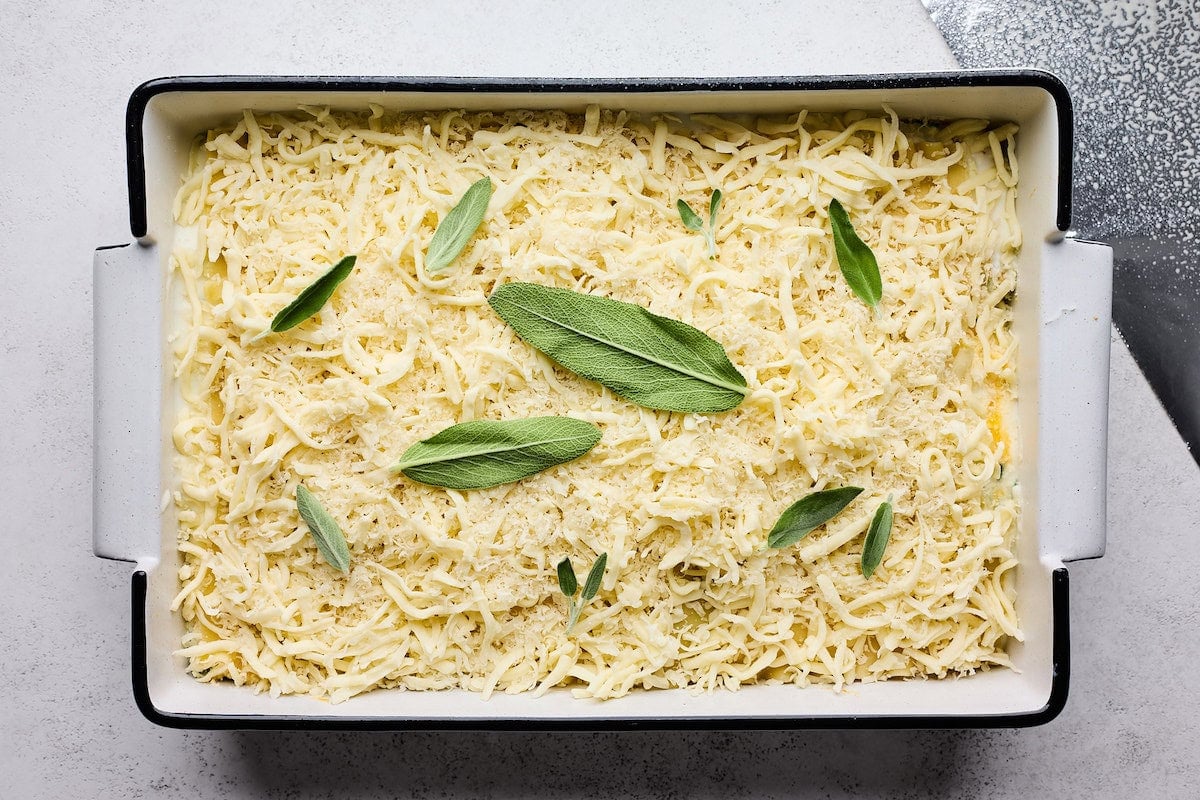 butternut squash being assembled in baking dish with cheese and sage leaves on top.