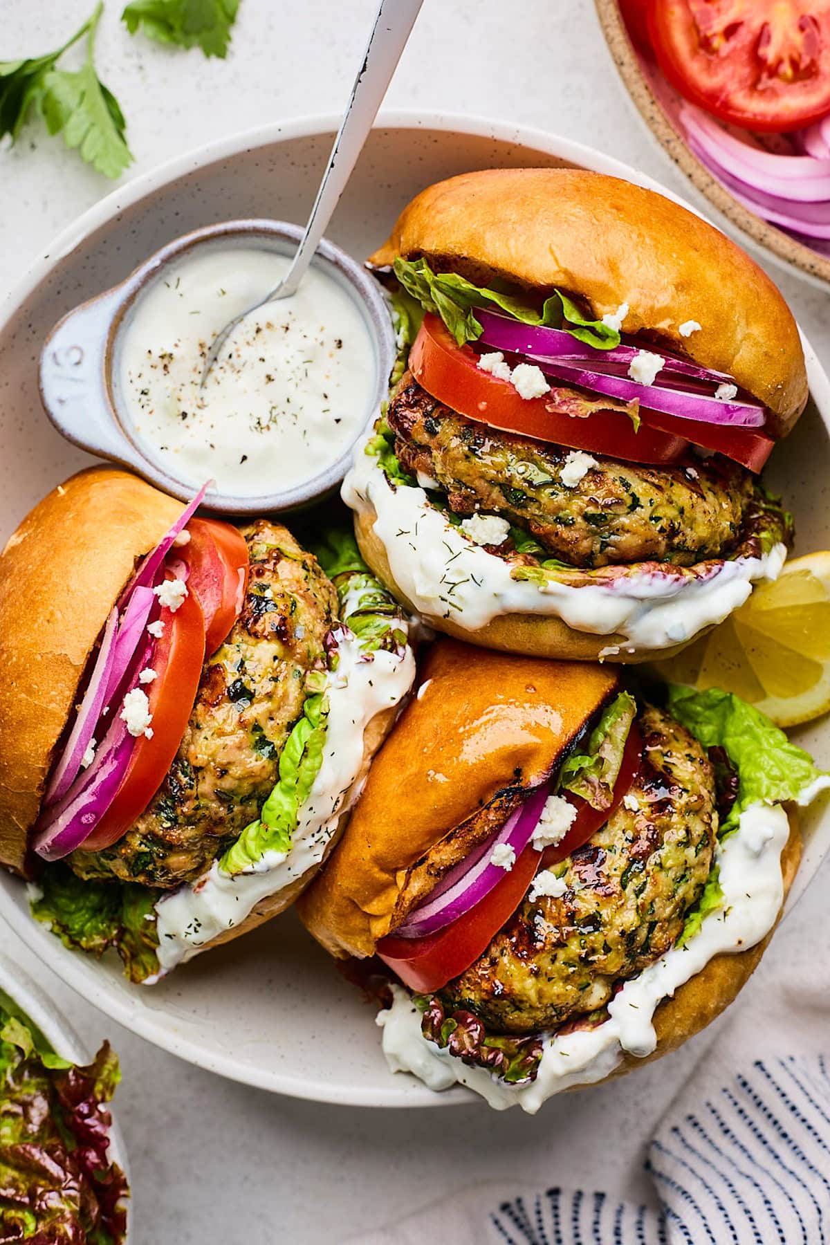 Three Greek turkey burgers on buns with red onion, tomato, lettuce, and tzatziki sauce on plate.