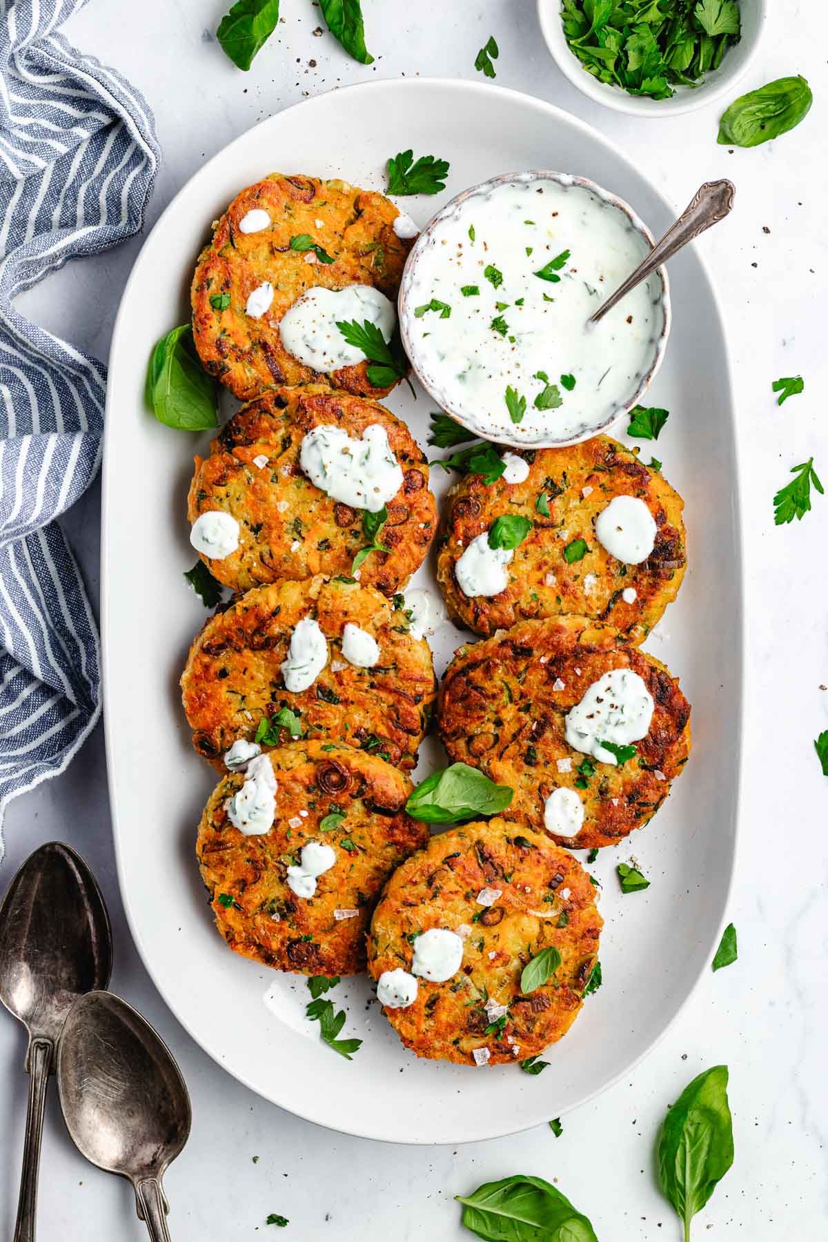 vegetable chickpea fritters on platter with lemon herb yogurt sauce and fresh herbs.
