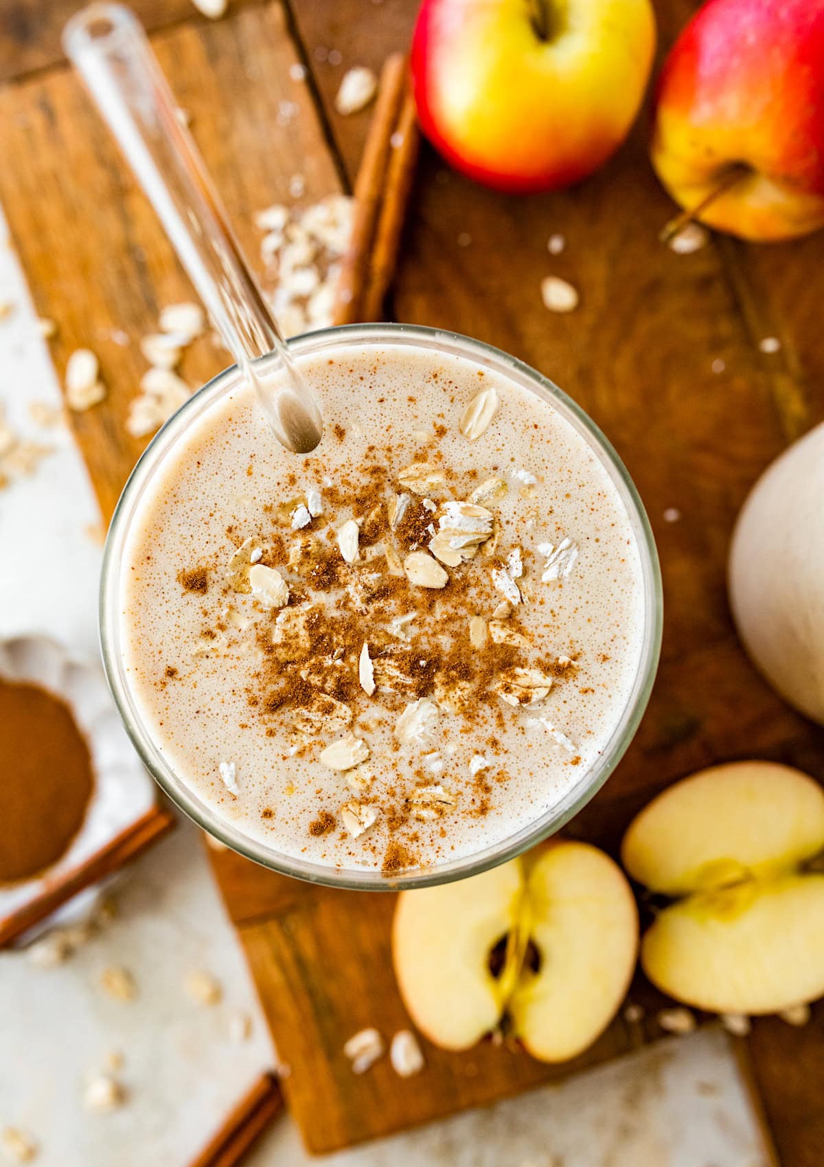 apple smoothie in glass with straw topped with cinnamon and oats with fresh apples on wood board.