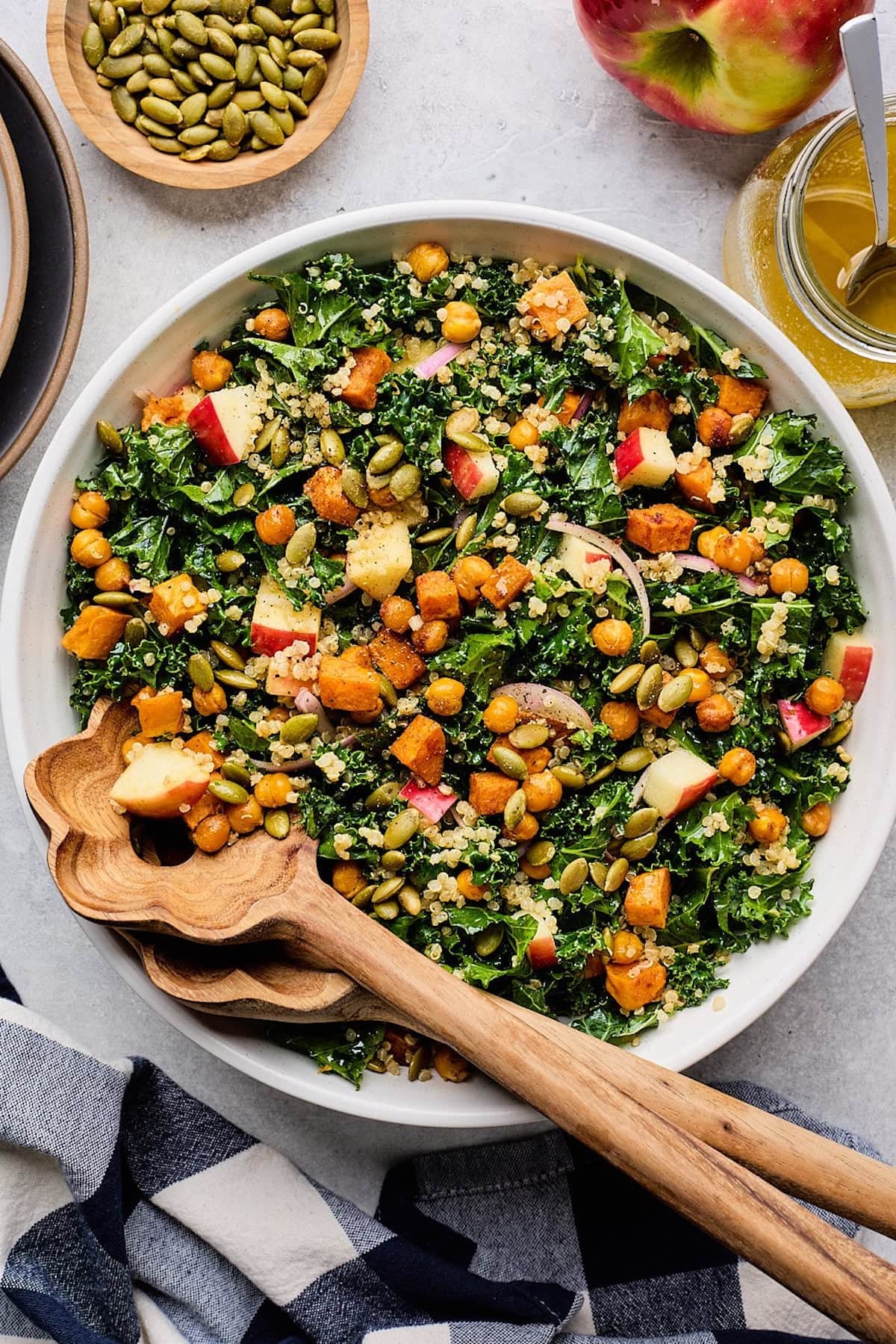 sweet potato kale salad with crispy chickpeas, quinoa, apples, pepitas, and red onion in bowl with wood salad servers.