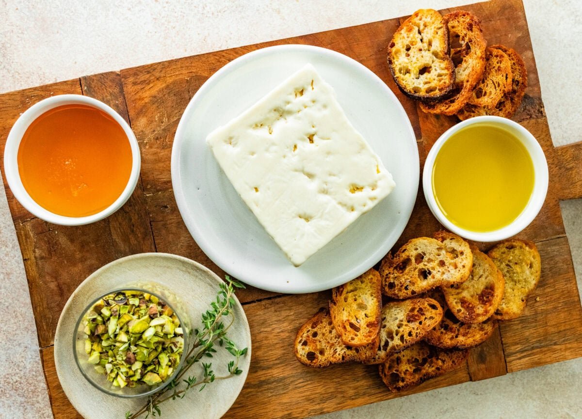 block of feta cheese, olive oil, honey, pistachios, and time on wood board with crostini to make baked feta.
