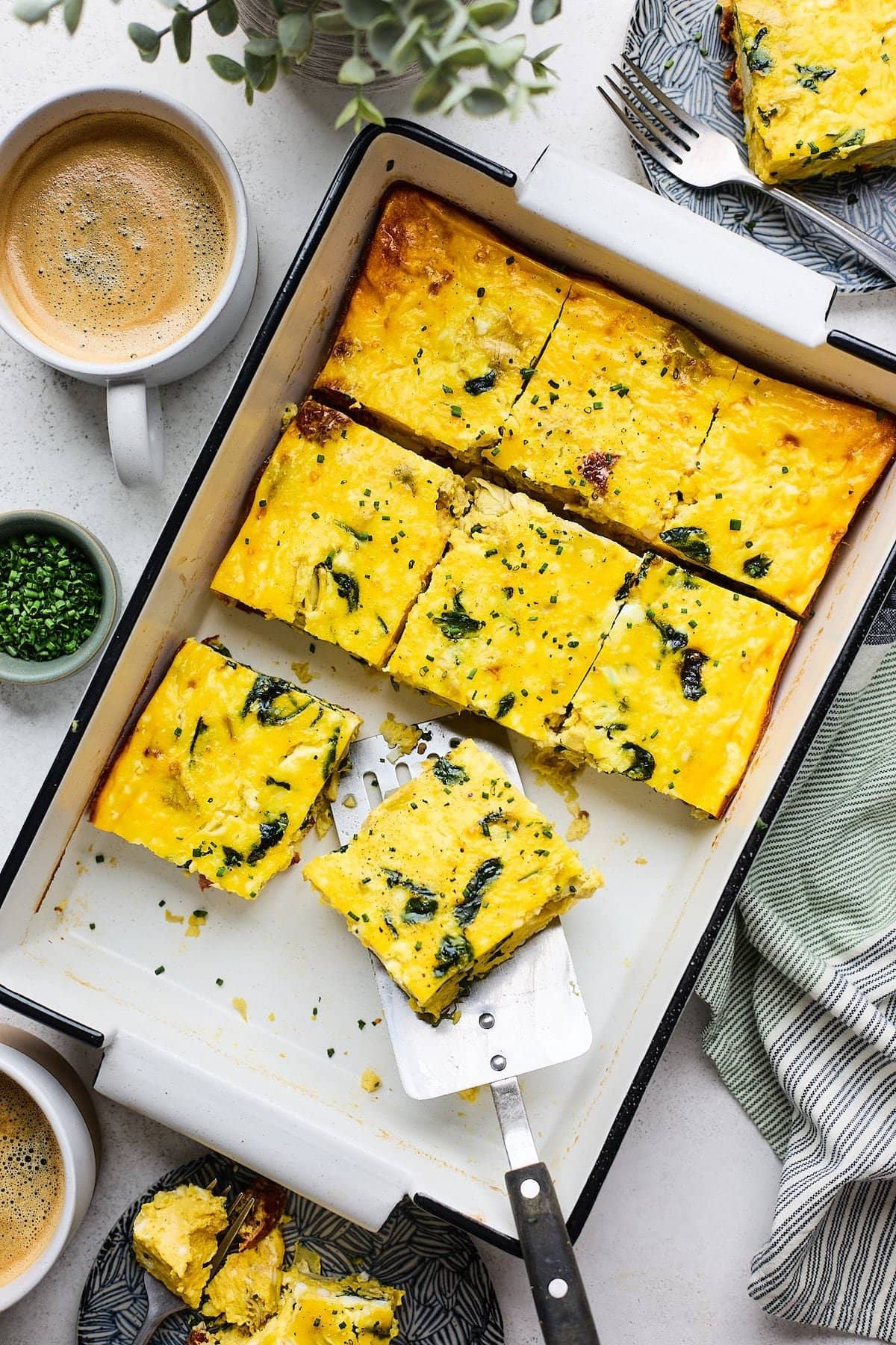 Spinach Artichoke Sun-Dried Tomato Breakfast Casserole cut into squares and being served with metal spatula in white baking dish.