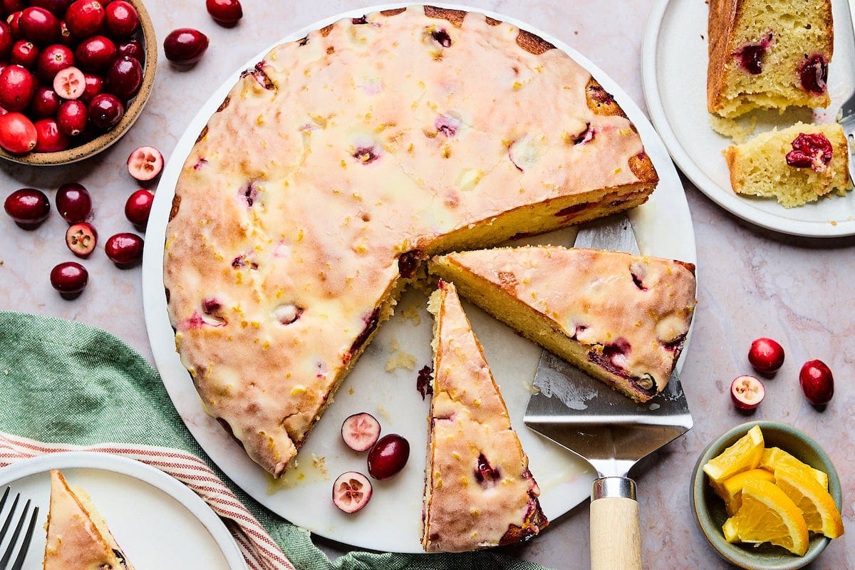 cranberry orange cake with orange glaze being cut into pieces on marble board.