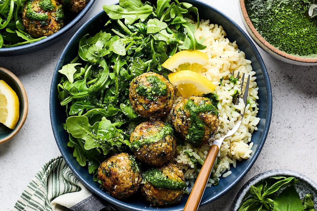 lemon herb meatballs in bowl with arugula, rice, basil vinaigrette, lemon slices, and fork.