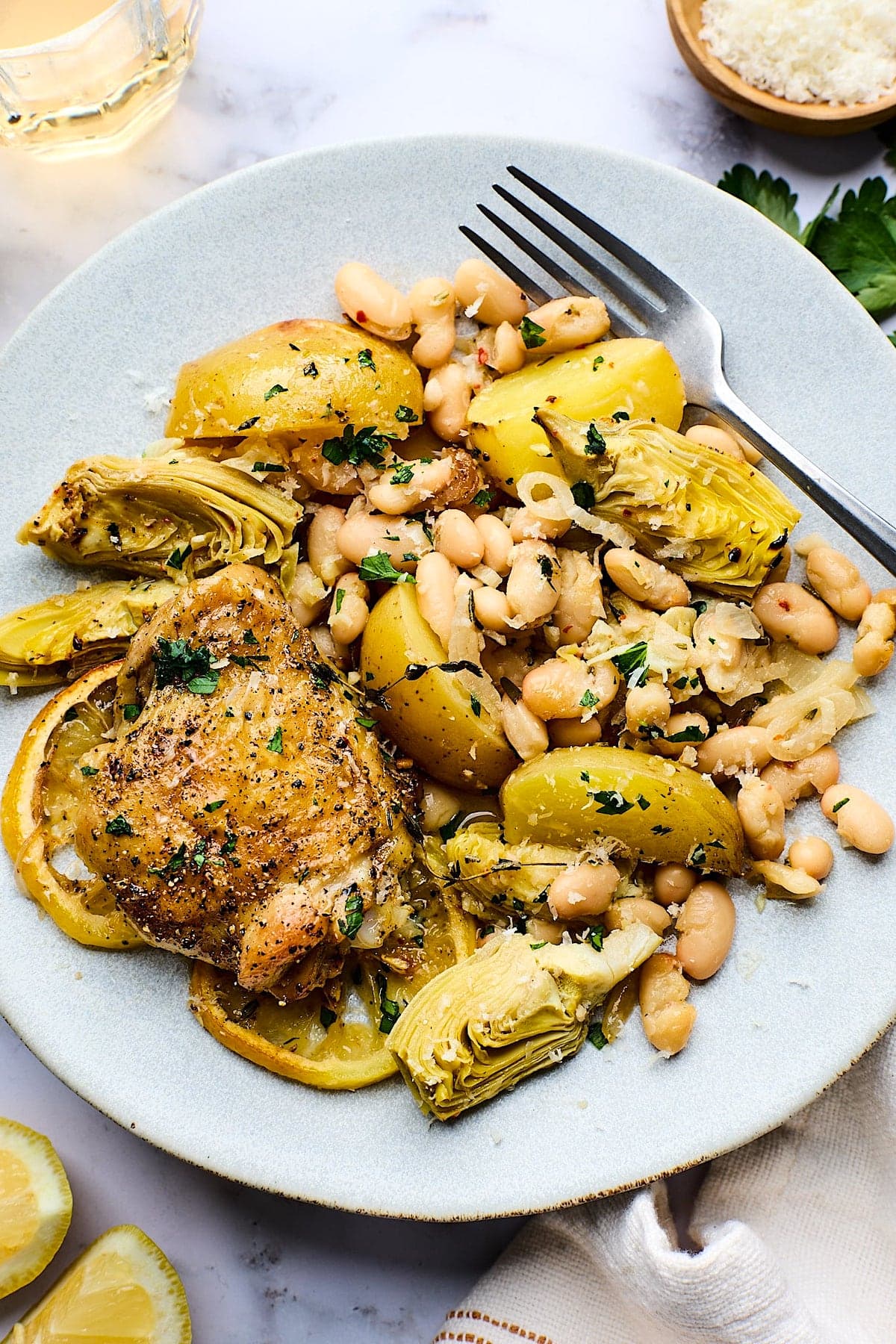 oven roasted chicken with potatoes, white beans, and artichokes on plate with herbs, parmesan cheese, and fork on plate. 
