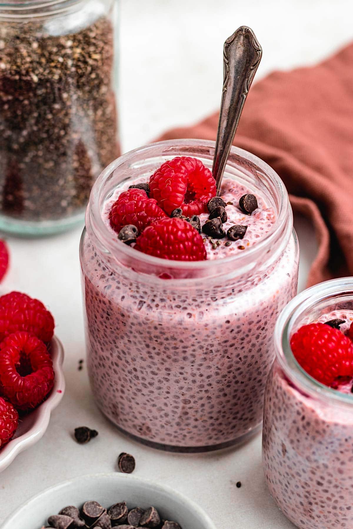 raspberry chia pudding in a jar witha spoon and topped with fresh raspberries and chocolate chips.