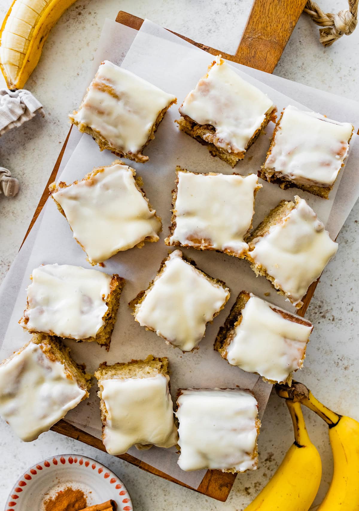 cinnamon swirl banana cake cut into squares with cream cheese glaze on wood board with parchment paper.