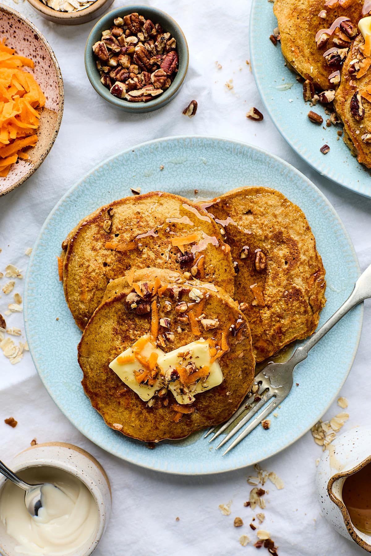 three carrot cake pancakes with butter, pecans, and syrup on plate with fork.