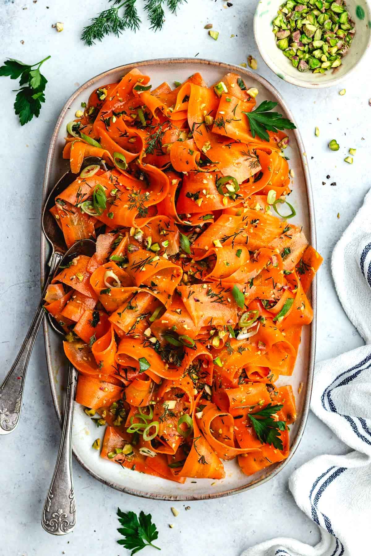 carrot ribbon salad with green onions, herbs, and pistachios on platter with serving spoons.