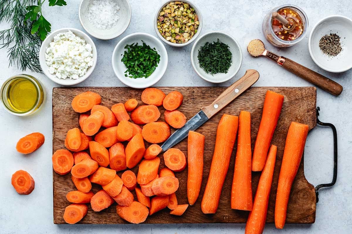 carrots being chopped on cutting board with knife and ingredients in bowls to make roasted carrots with hot honey, pistachios, and feta.