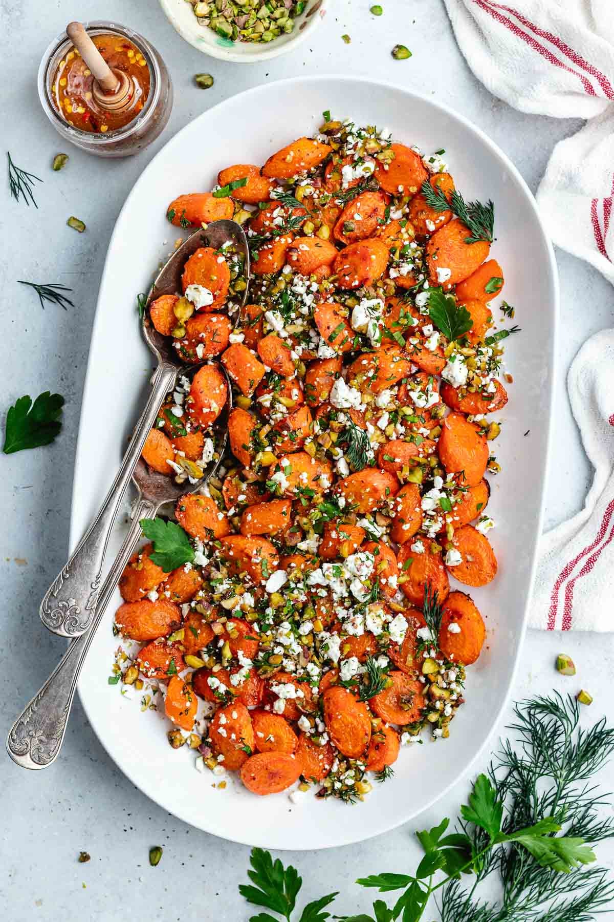 roasted carrots with pistachios, feta, herbs, and hot honey on platter with serving spoons.