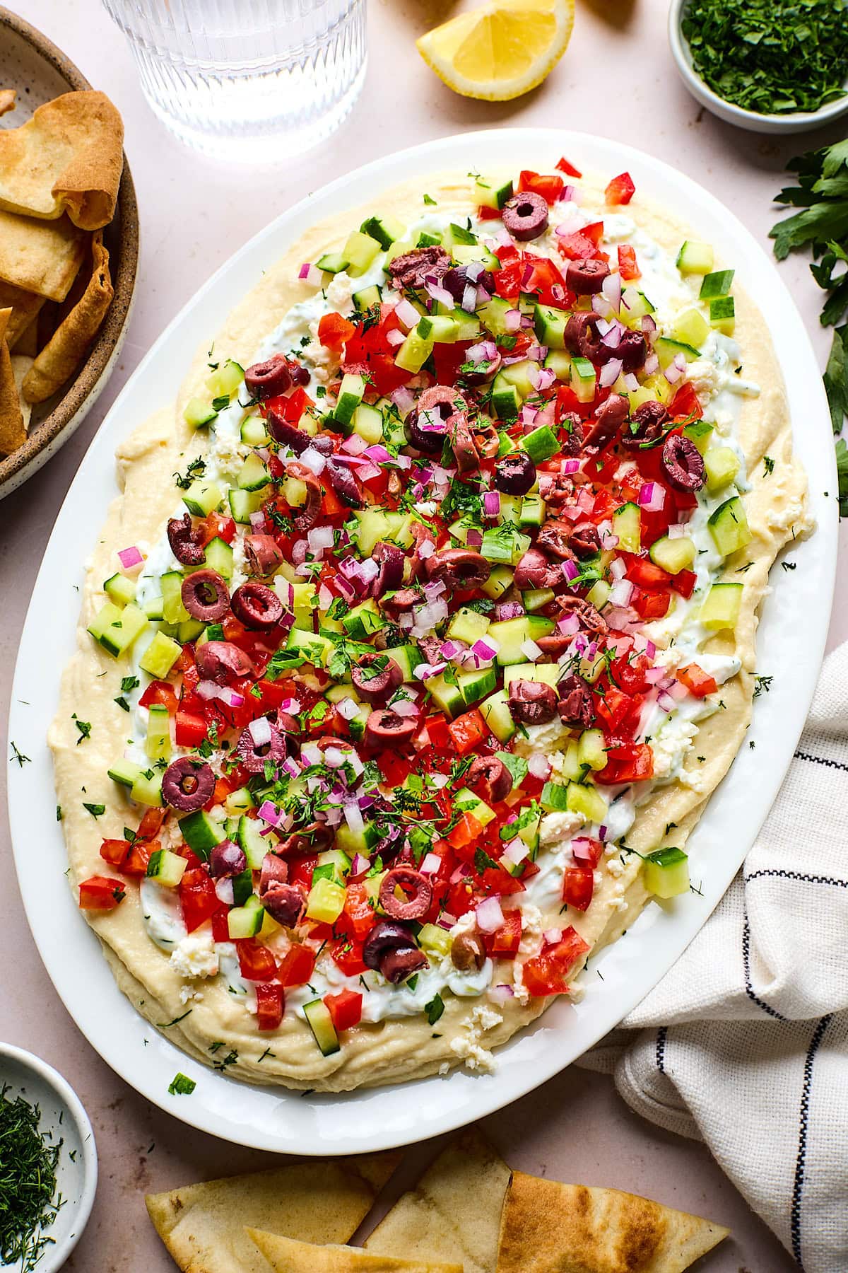 Greek layered dip with hummus, tzatziki, feta, cucumber, tomatoes, red onion, olives, and herbs on platter.